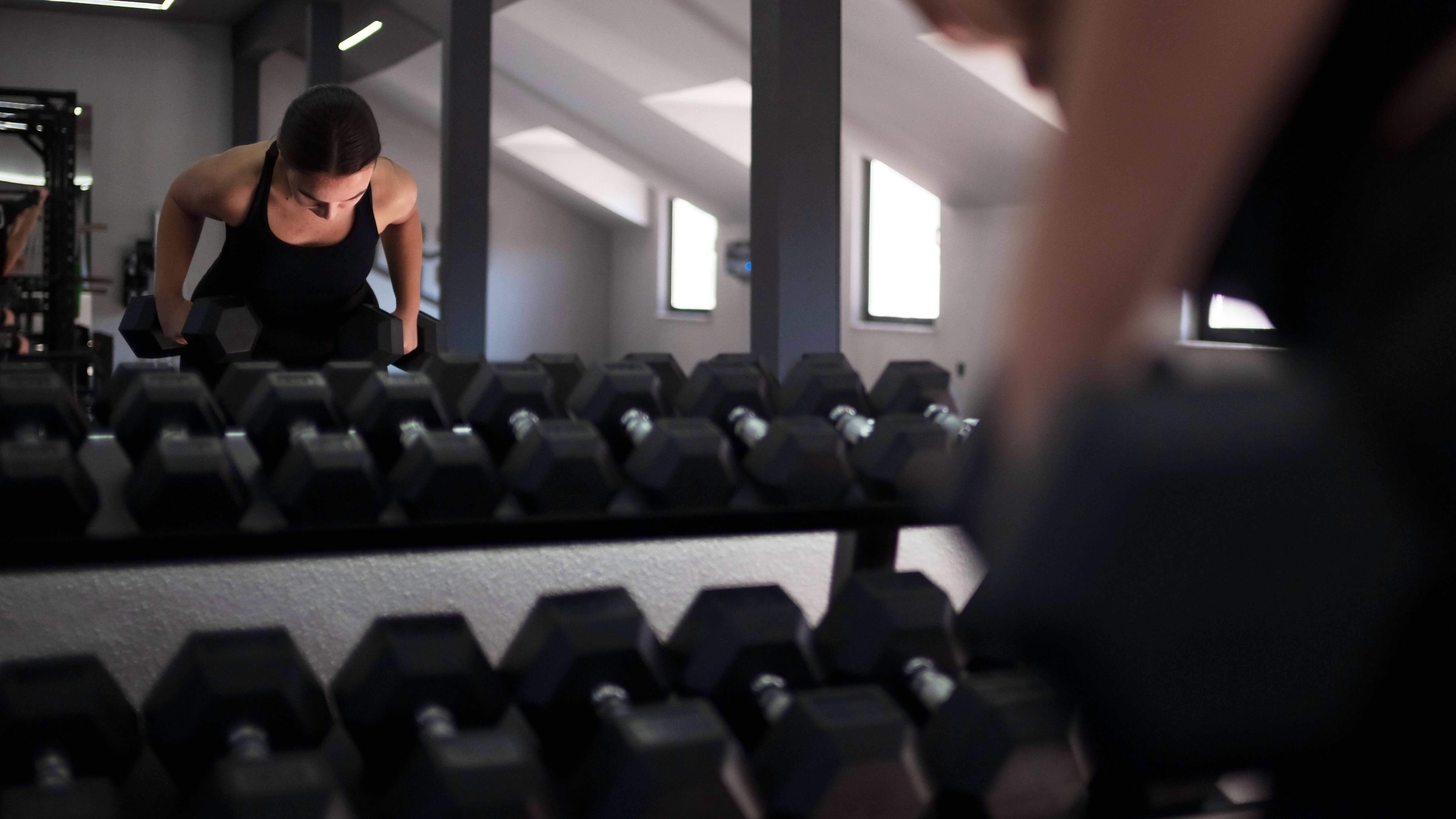 Eine Frau trainiert in einem Fitnessstudio mit Kurzhanteln. Sie sieht sich im Spiegel. Davor steht ein Regal mit weiteren Kurzhanteln.