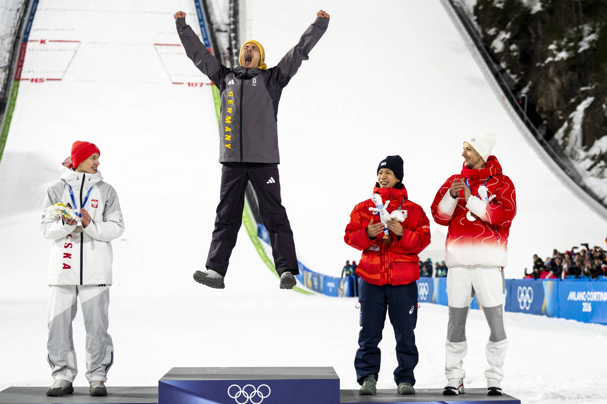 Philipp Raimund hat Gold beim Skispringen gewonnen. Der Sportler steht bei der Siegerehrung auf dem Treppchen und springt hoch in die Luft.