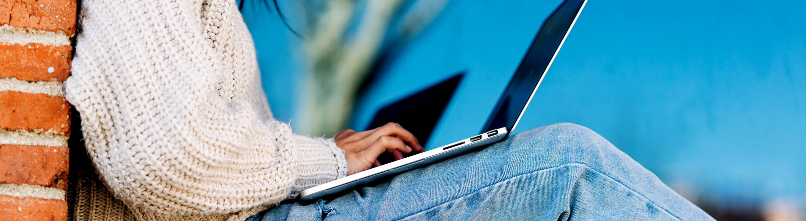 Eine Frau lehnt an einer Hauswand balanciert ihren Laptop auf ihren Knien und tippt auf der Tastatur