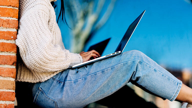 Eine Frau lehnt an einer Hauswand balanciert ihren Laptop auf ihren Knien und tippt auf der Tastatur