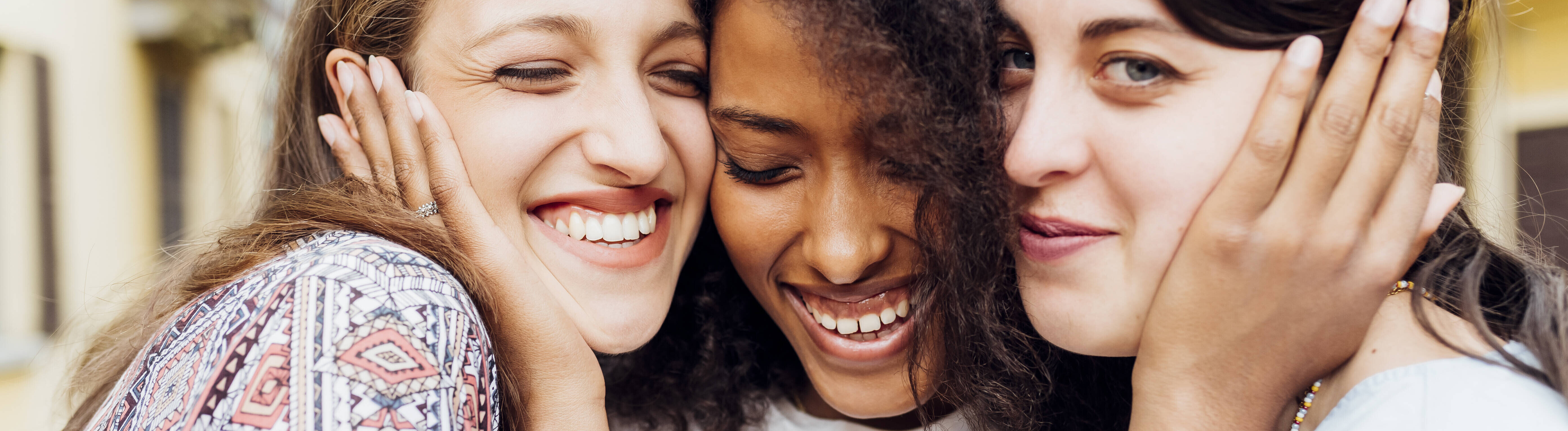 Drei Frauen halten ihre Gesicht innig nebeneinander. Die Frau in der Mitte hält ihre Hände an das Gesicht der beiden anderen Frauen. Sie lachen.