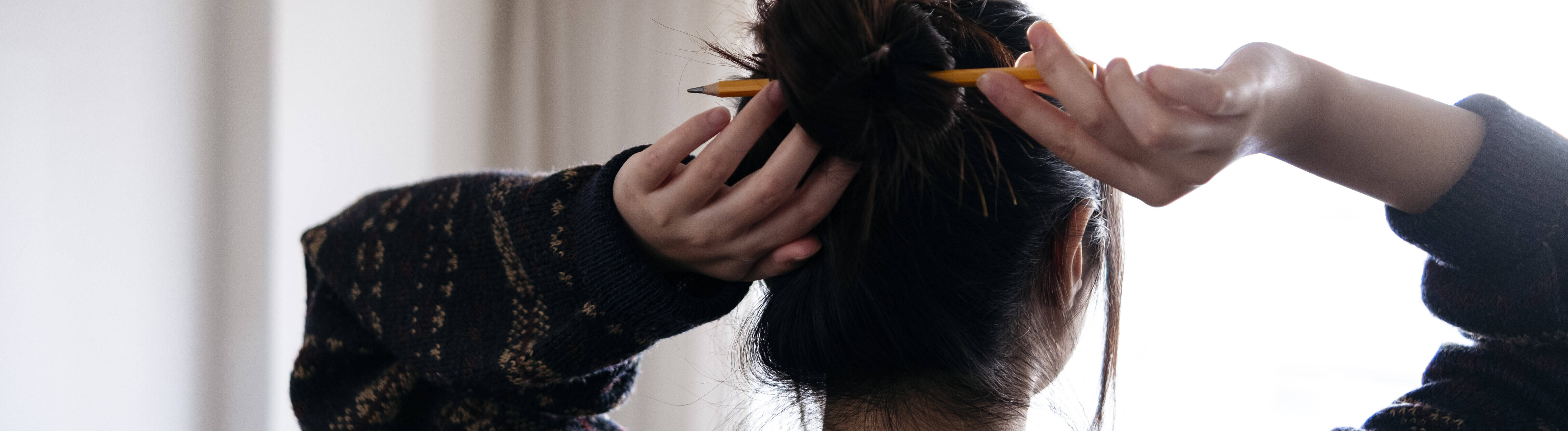 Eine junge Frau steht mit dem Rücke zur Kamera vor einem Fenster im Wohnzimmer. Sie macht sich die Haare mit einem Bleistift zu einem Dutt zusammen.