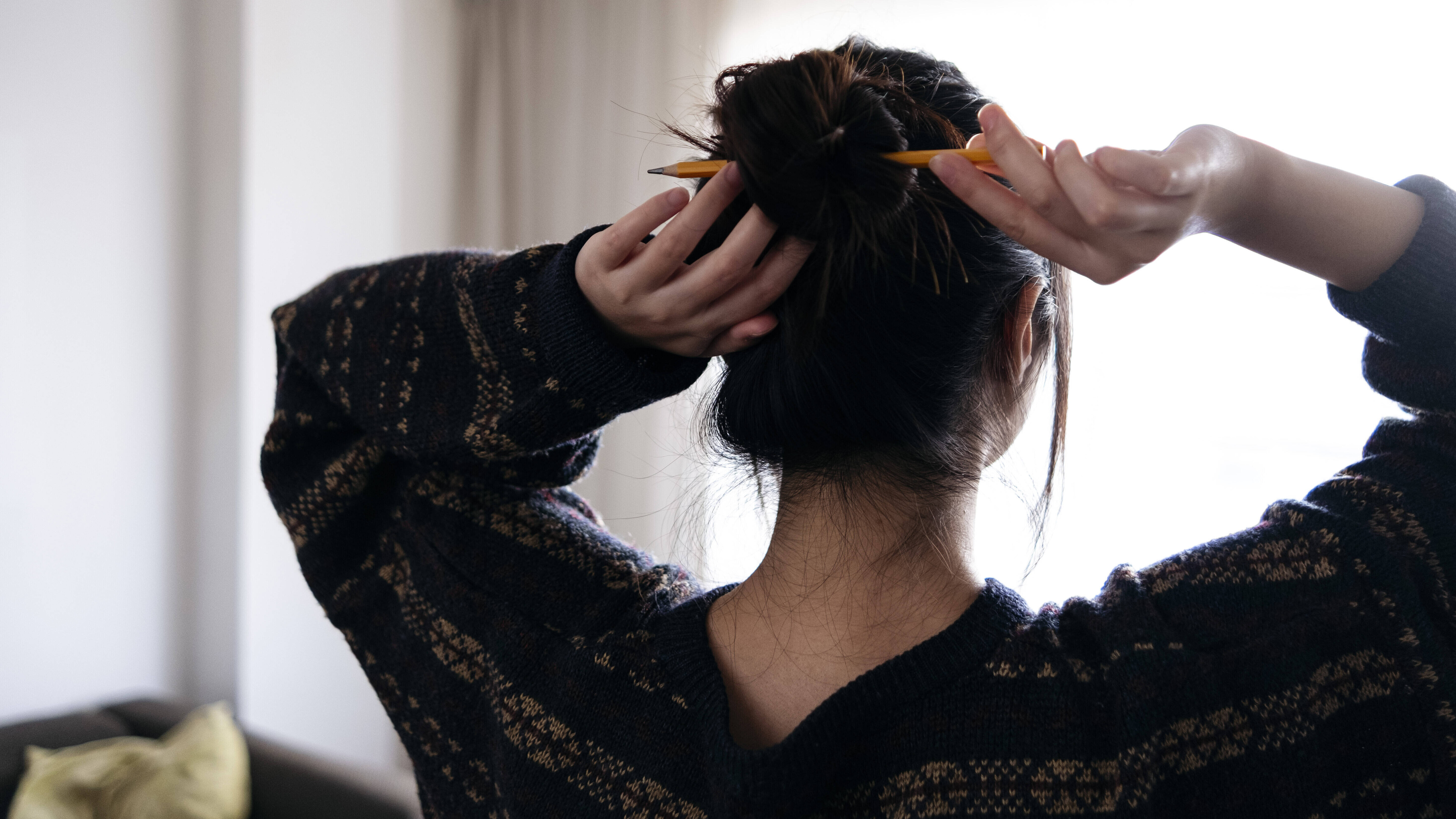 Eine junge Frau steht mit dem Rücke zur Kamera vor einem Fenster im Wohnzimmer. Sie macht sich die Haare mit einem Bleistift zu einem Dutt zusammen.