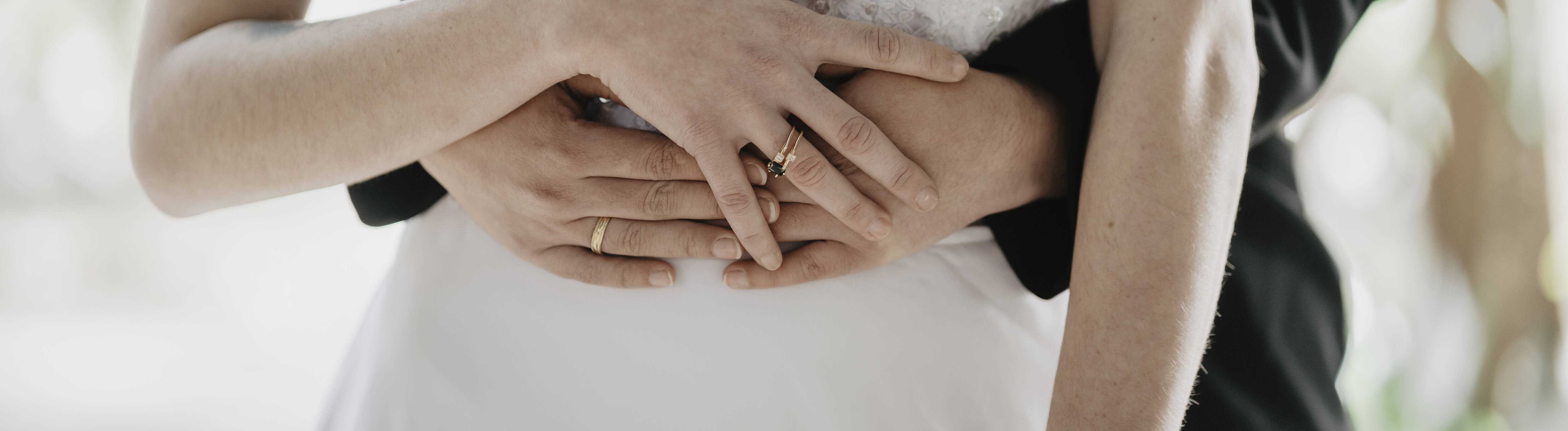Ein Hochzeitspaar steht hintereinander. Eine der beiden Frauen trägt ein weißes Hochzeitskleid. Die andere Frau trägt einen schwarzen Anzug. Sie umarmen sich, dabei halten sie ihre Hände übereinander.