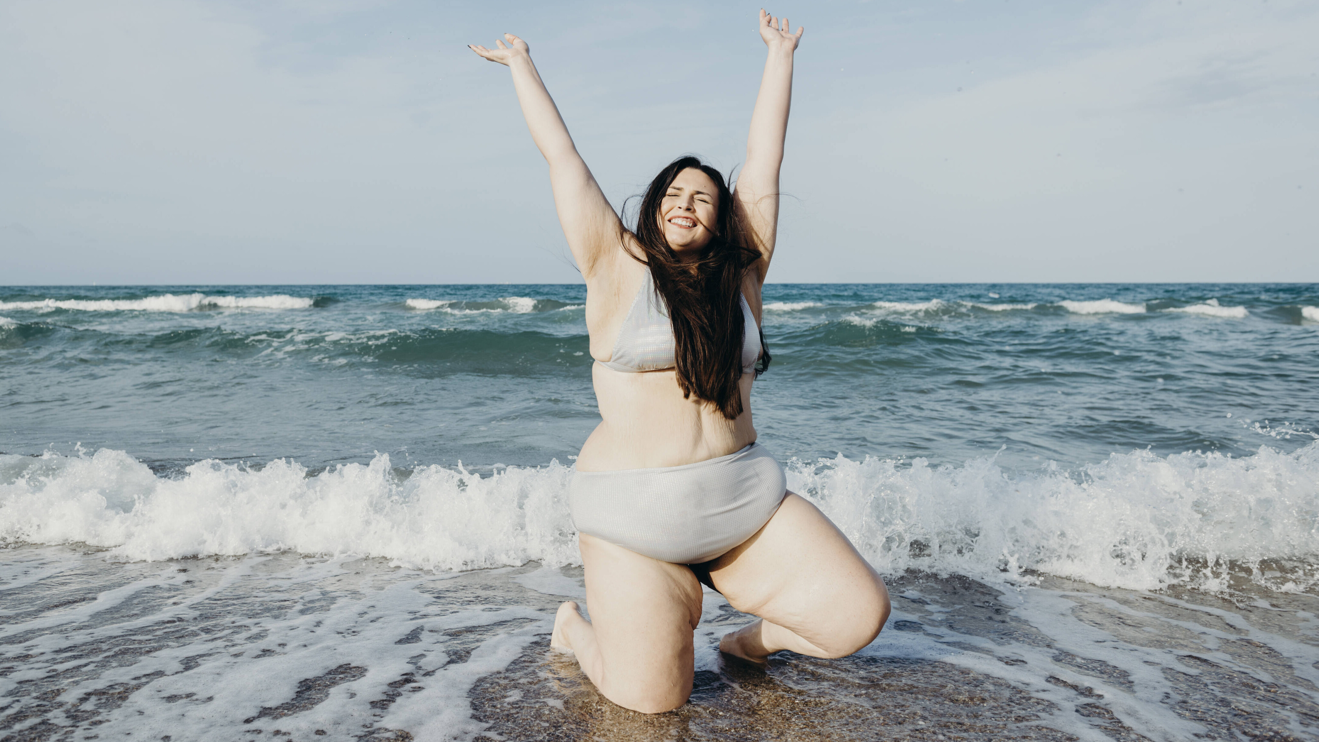Eine Frau im Bikini kniet an einem Strand im Wasser und hebt ihre Arme in die Luft. Sie lächelt und hat ihre Augen geschlossen.