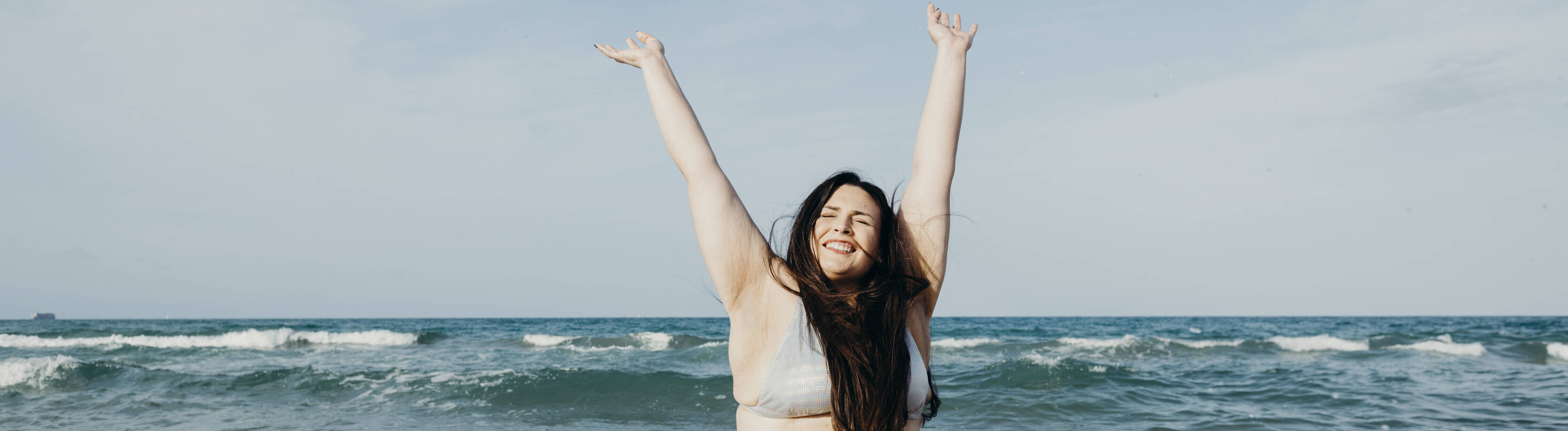 Eine Frau im Bikini kniet an einem Strand im Wasser und hebt ihre Arme in die Luft. Sie lächelt und hat ihre Augen geschlossen.