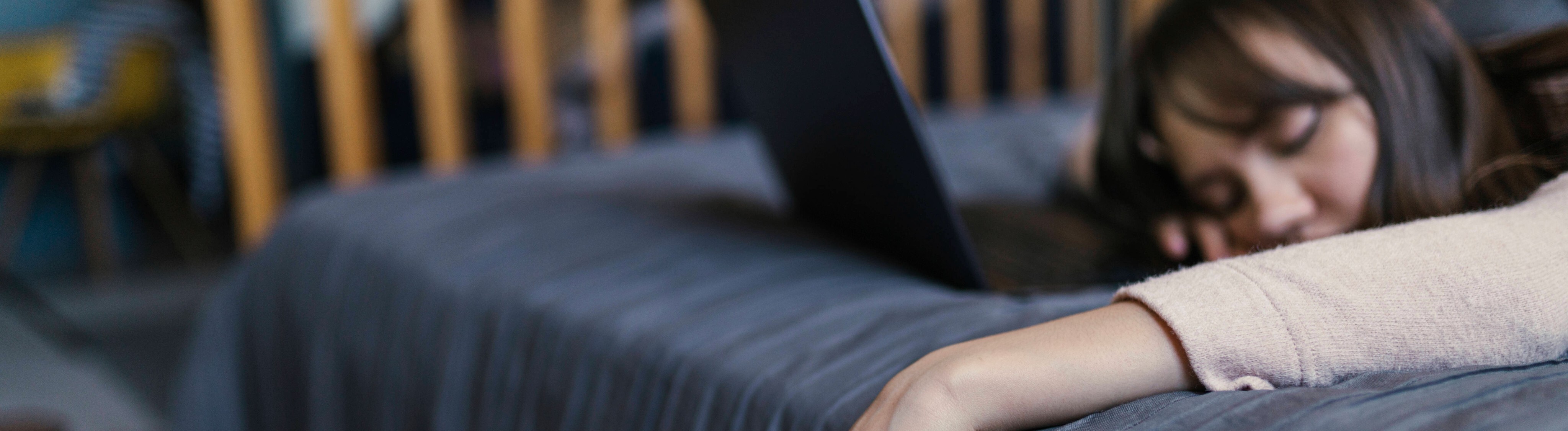 Eine Frau liegt auf dem Bett, die Hand, in der sie ein Smartphone hält, hängt schlaff herab, Vor ihr ein Laptop.