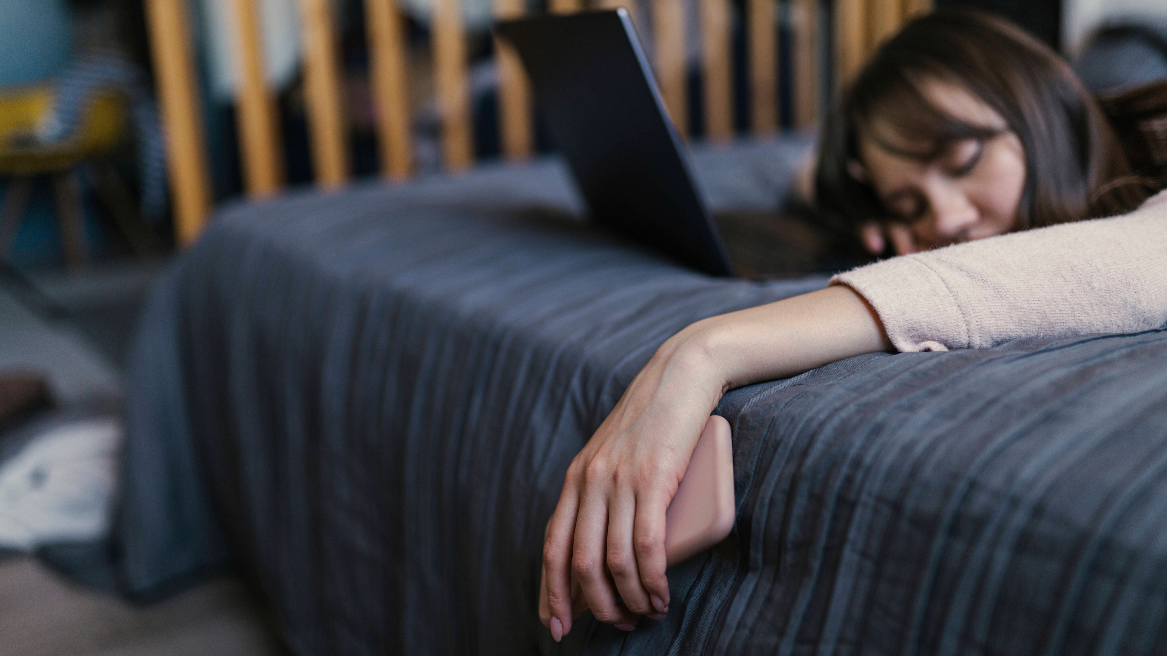 Eine Frau liegt auf dem Bett, die Hand, in der sie ein Smartphone hält, hängt schlaff herab, Vor ihr ein Laptop.