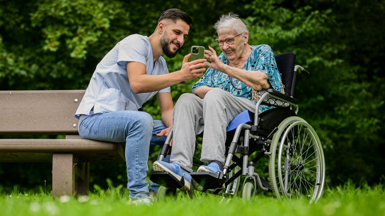 Mann sitzt auf Parkbank und schaut mit alter Frau im Rollstuhl auf Smartphone