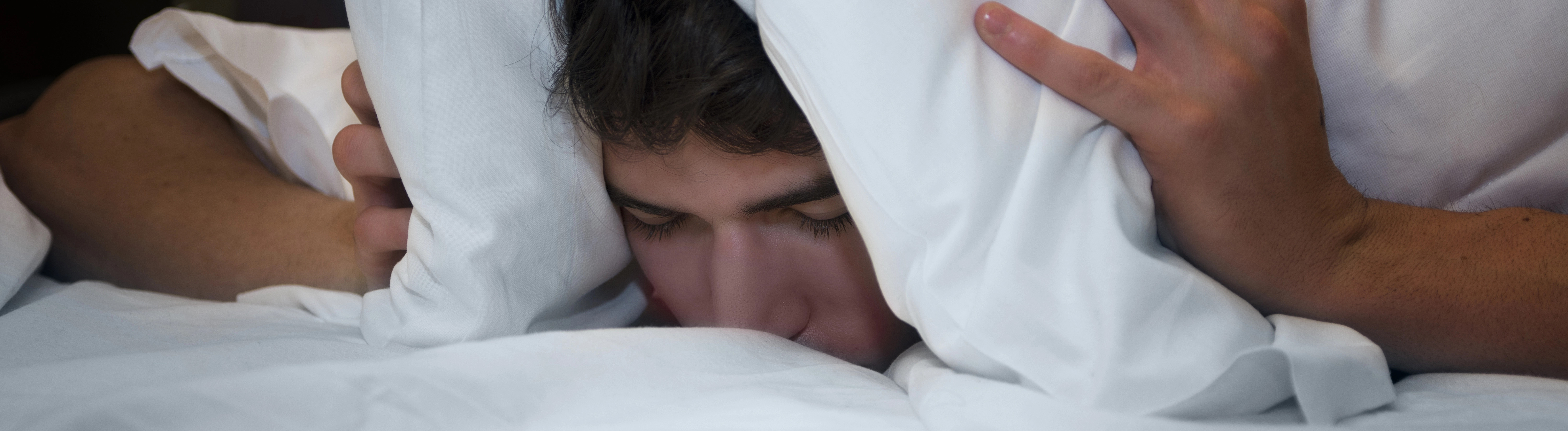 Ein Mann zieht sich das Kopfkissen über den Kopf und die Ohren, vermutlich um sich vor Lärm zu schützen.