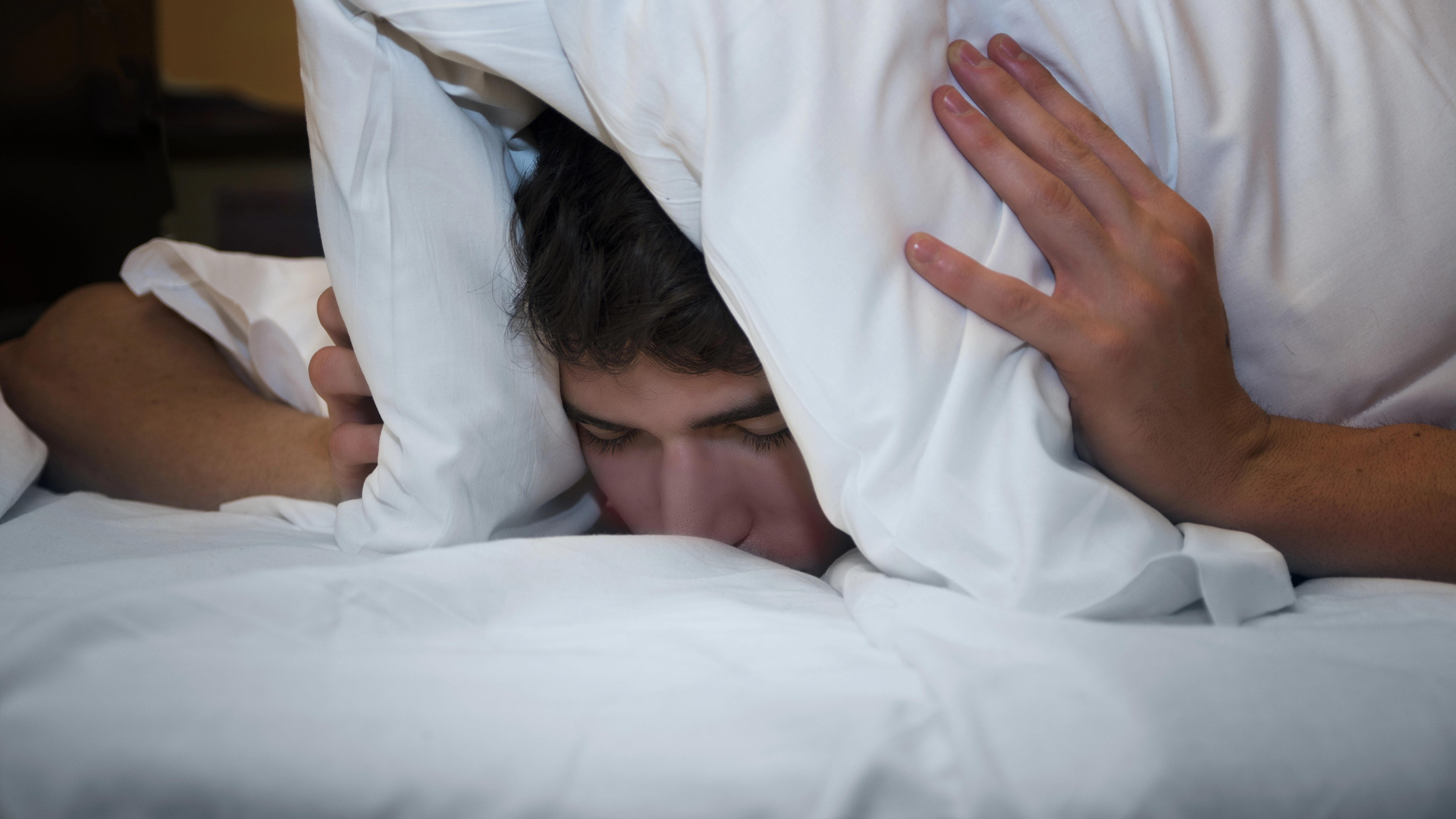 Ein Mann zieht sich das Kopfkissen über den Kopf und die Ohren, vermutlich um sich vor Lärm zu schützen.