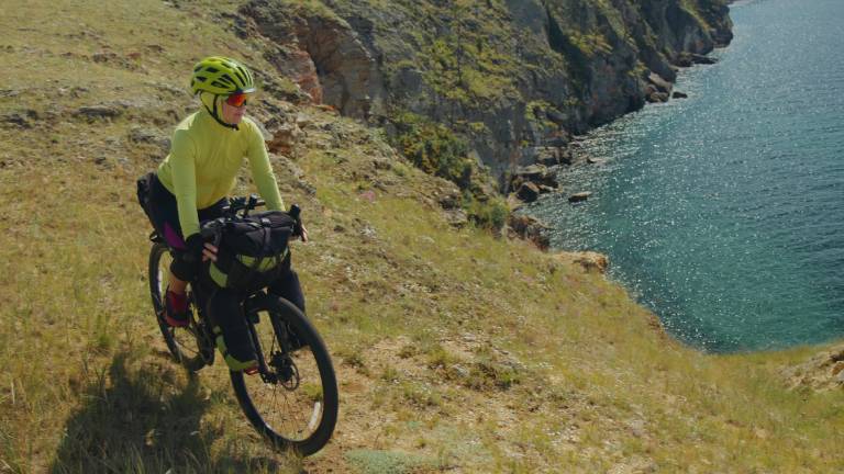 Eine Frau fährt mit ihrem Fahrrad an einer Steilküste entlang. Sie trägt neonfarbene Fahrradkleidung und einen Helm. Ihr Rad ist mit Satteltaschen bepackt. Im Hintergrund sieht man das Meer.