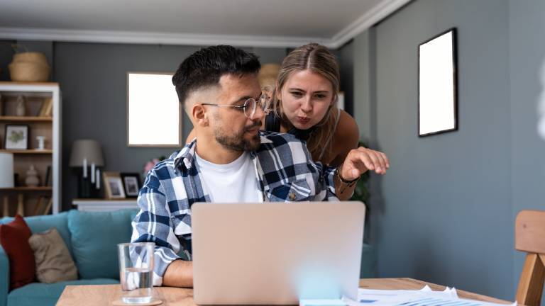 Eine Frau lenkt ihren Mann ab, der im Homeoffice in der Küche vor Notizen und seinem Laptop sitzt.