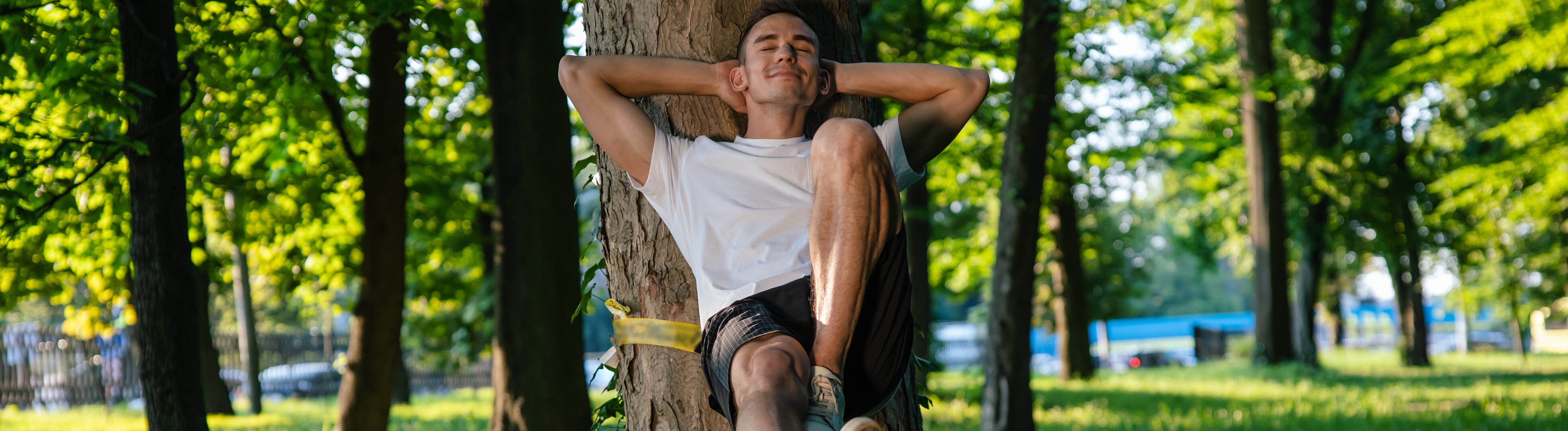 Ein Mann sitzt an einem Baum gelehnt auf einer Slackline und ruht sich zufrieden aus.