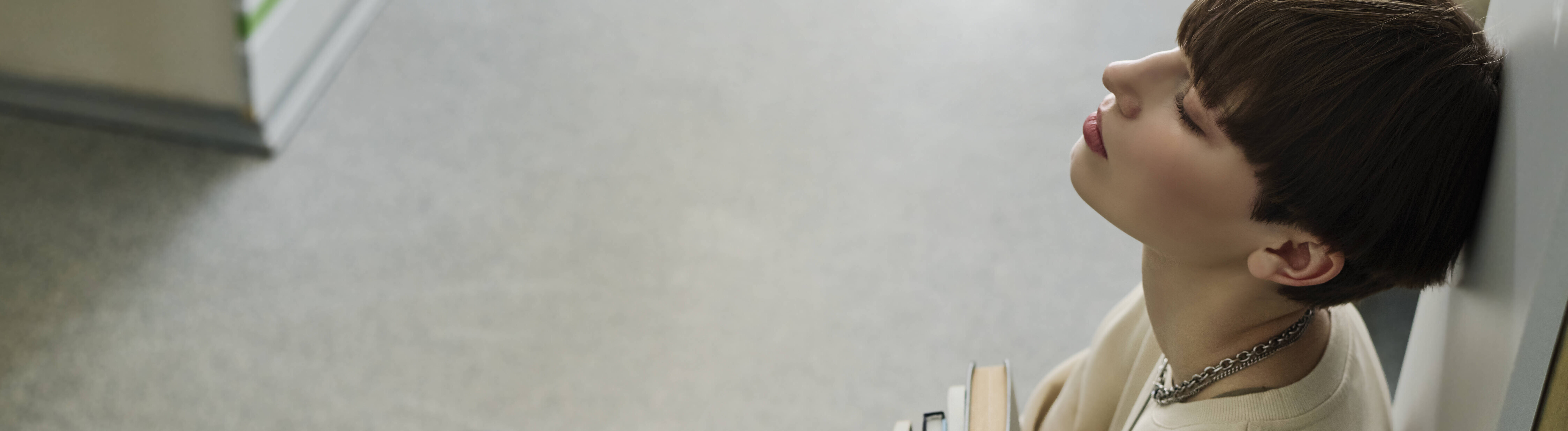 Ein Student sitzt in einer Bibliothek auf dem Boden und lehnt mit dem Rücken an ein Bücherregal an. Er hält Bücher in den Händen und hat die Augen geschlossen. Er wirkt erschöpft.
