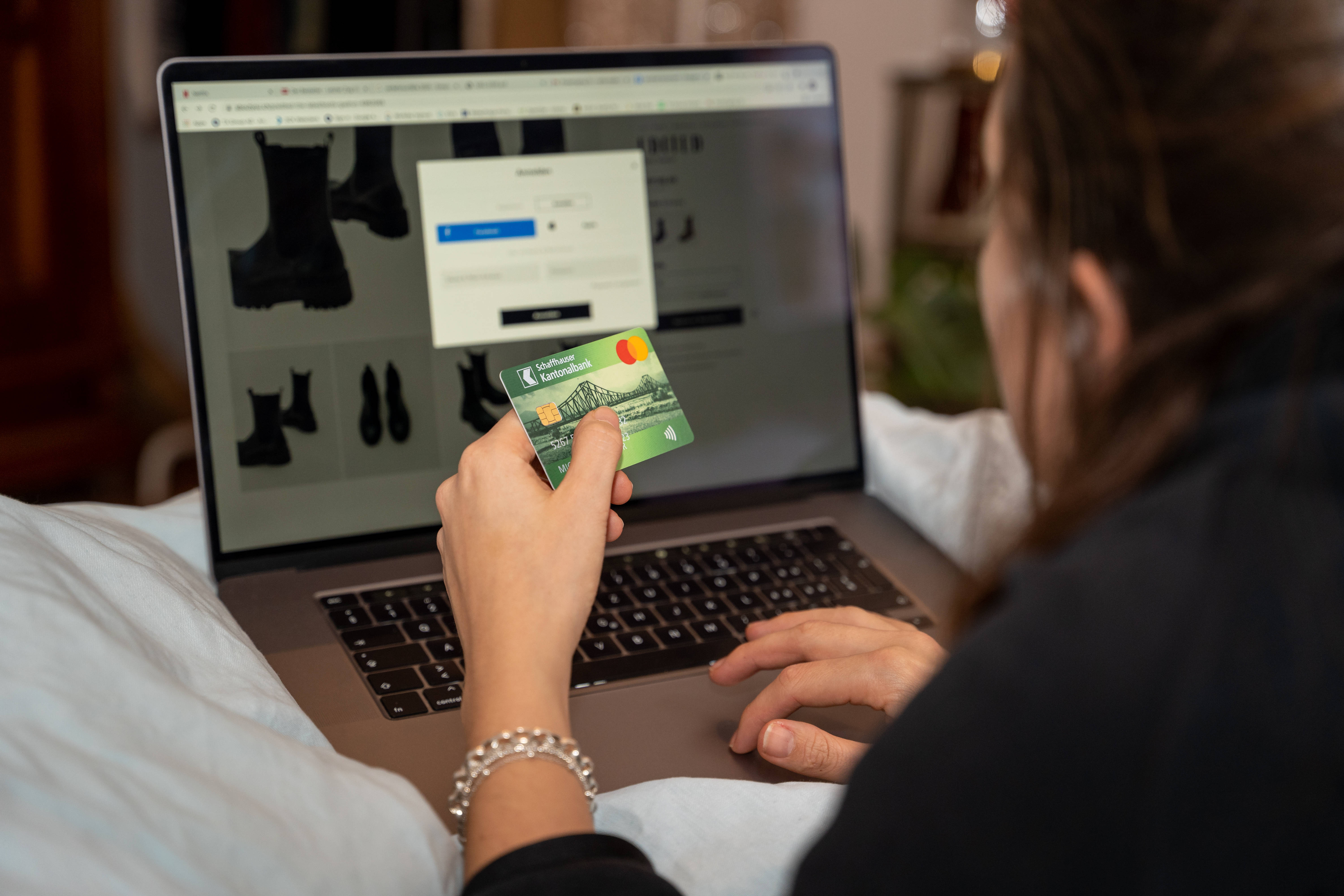 Eine Frau sitzt vor einem Laptop und hat eine Shoppingseite geöffnet. in der linken Hand hält sie ihre Kreditkarte, während sie mit der rechten tippt.