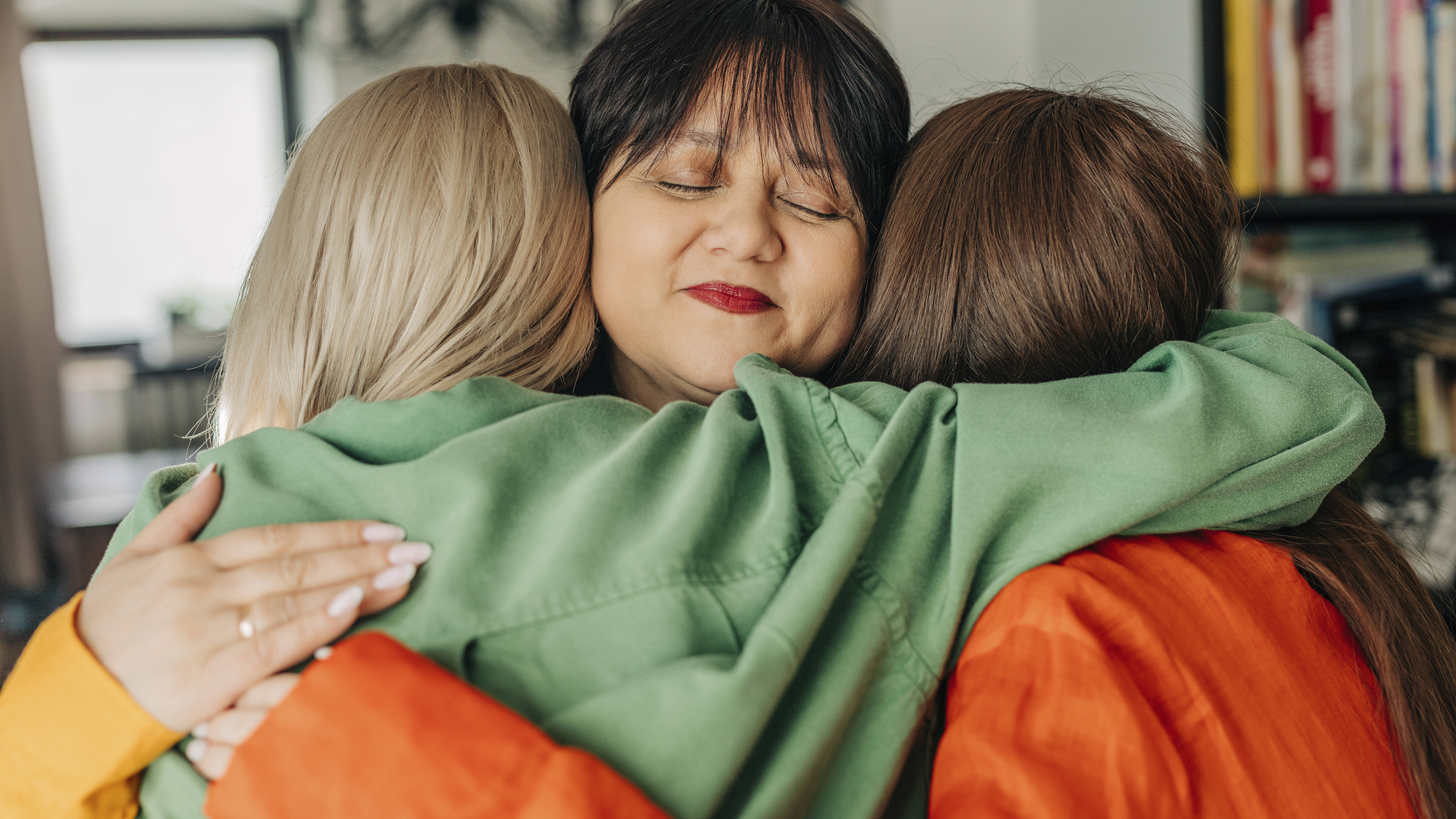 Zwei Frauen umarmen eine dritte Frau, die in der Mitte der beiden steht. Ihre Augen sind geschlossen und sie lächelt.