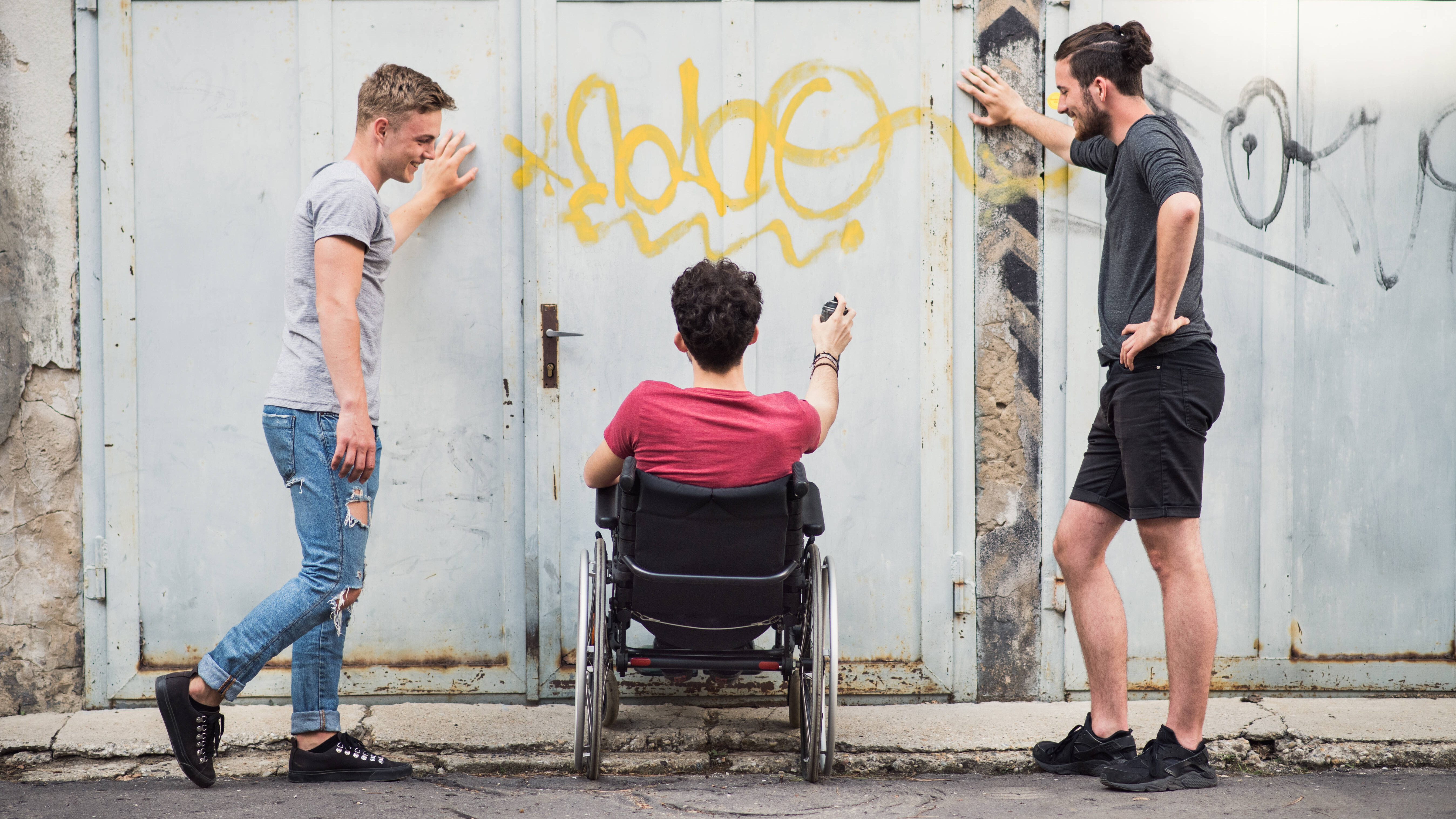 Drei Freunde beim Graffiti-Sprühen - zwei stehen, einer sitzt im Rollstuhl