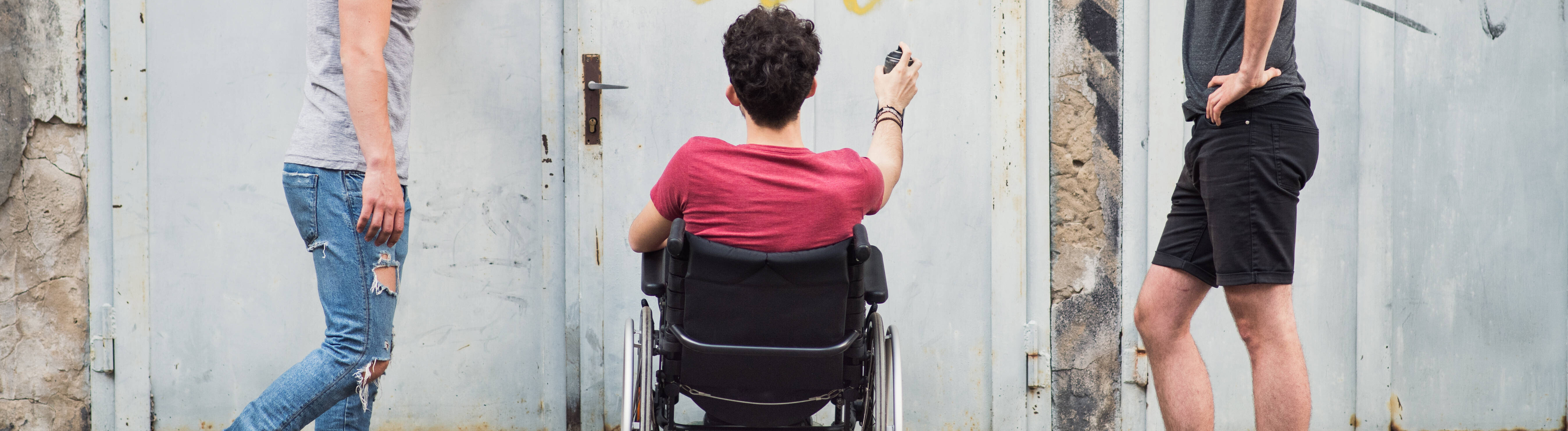 Drei Freunde beim Graffiti-Sprühen - zwei stehen, einer sitzt im Rollstuhl