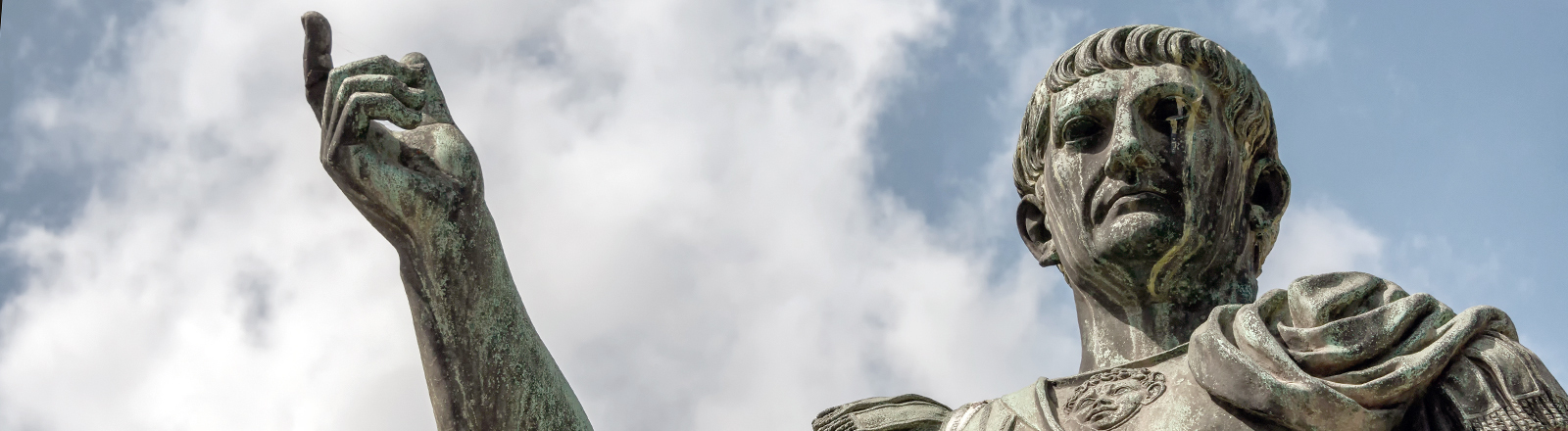 Untersichtige Fotografie einer Bronzestatue des römischen Kaisers Caesar Nervae Trajan vor weiß bewölktem blauen Himmel