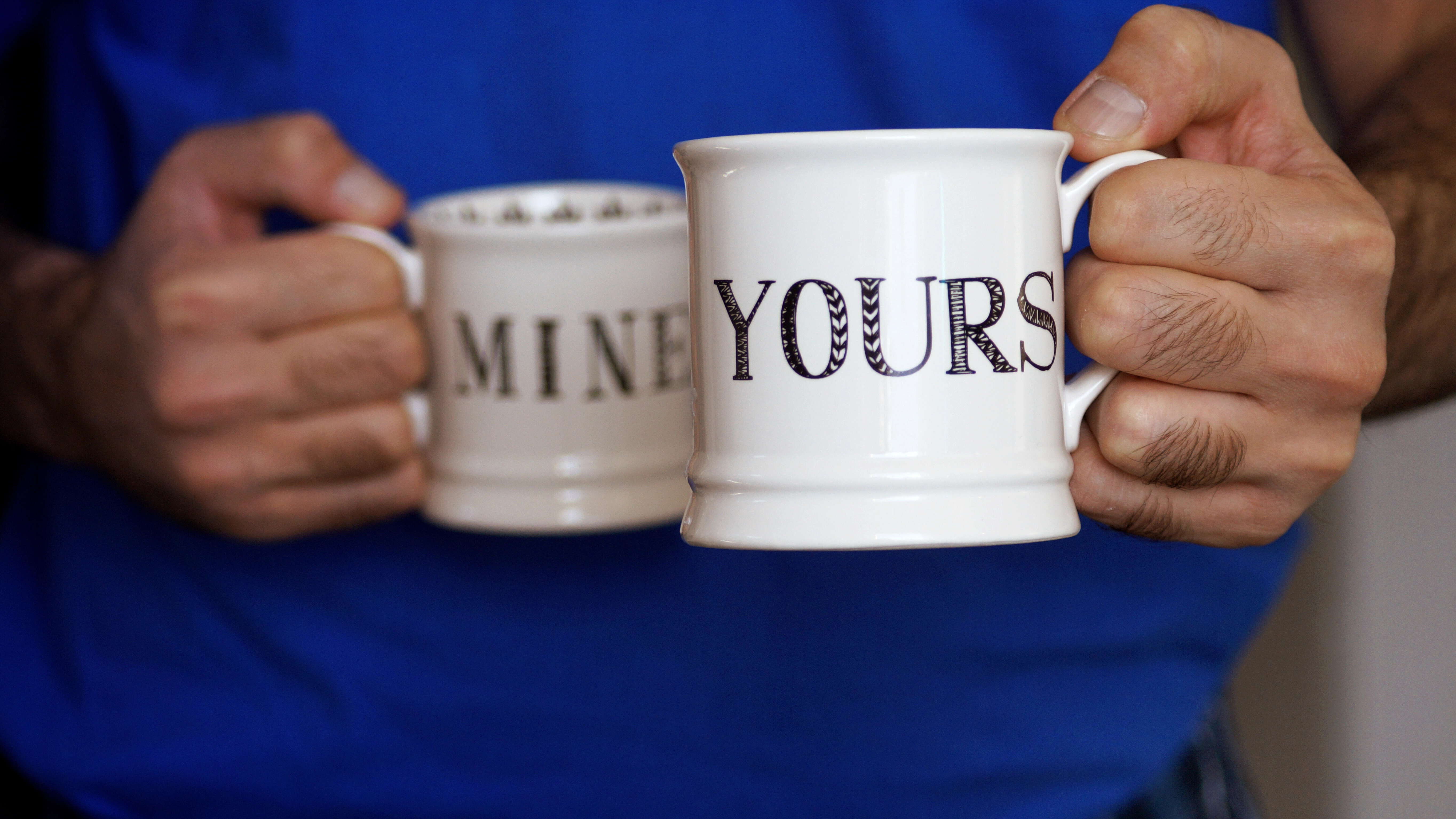 Ein Mann hält zwei Kaffeebecher in seinen Händen: Auf einem steht auf Englisch "mine", auf dem anderen "yours".