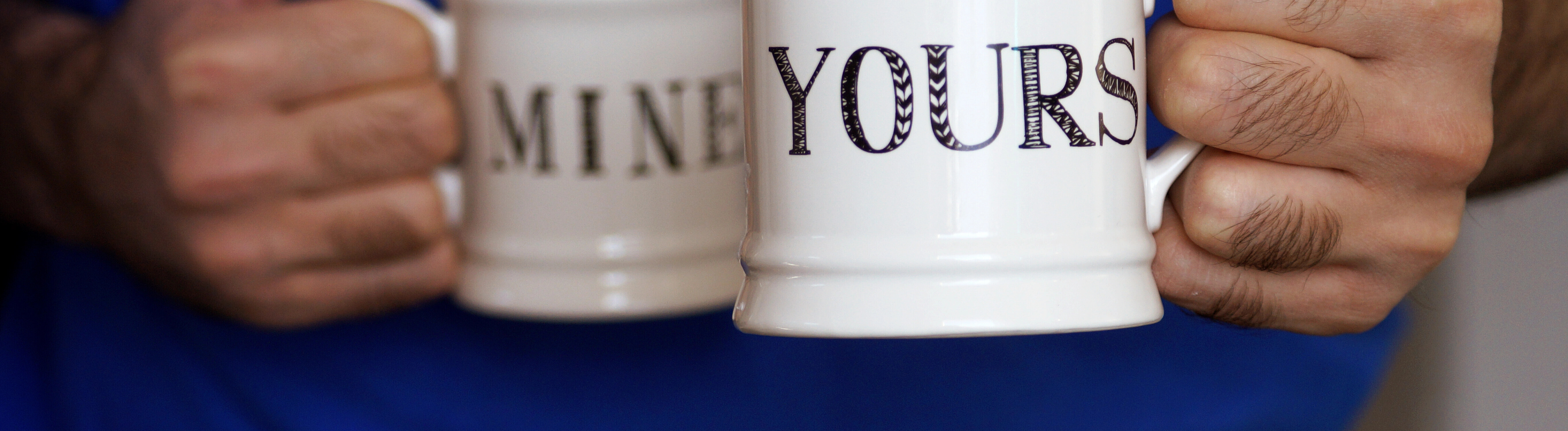 Ein Mann hält zwei Kaffeebecher in seinen Händen: Auf einem steht auf Englisch "mine", auf dem anderen "yours".