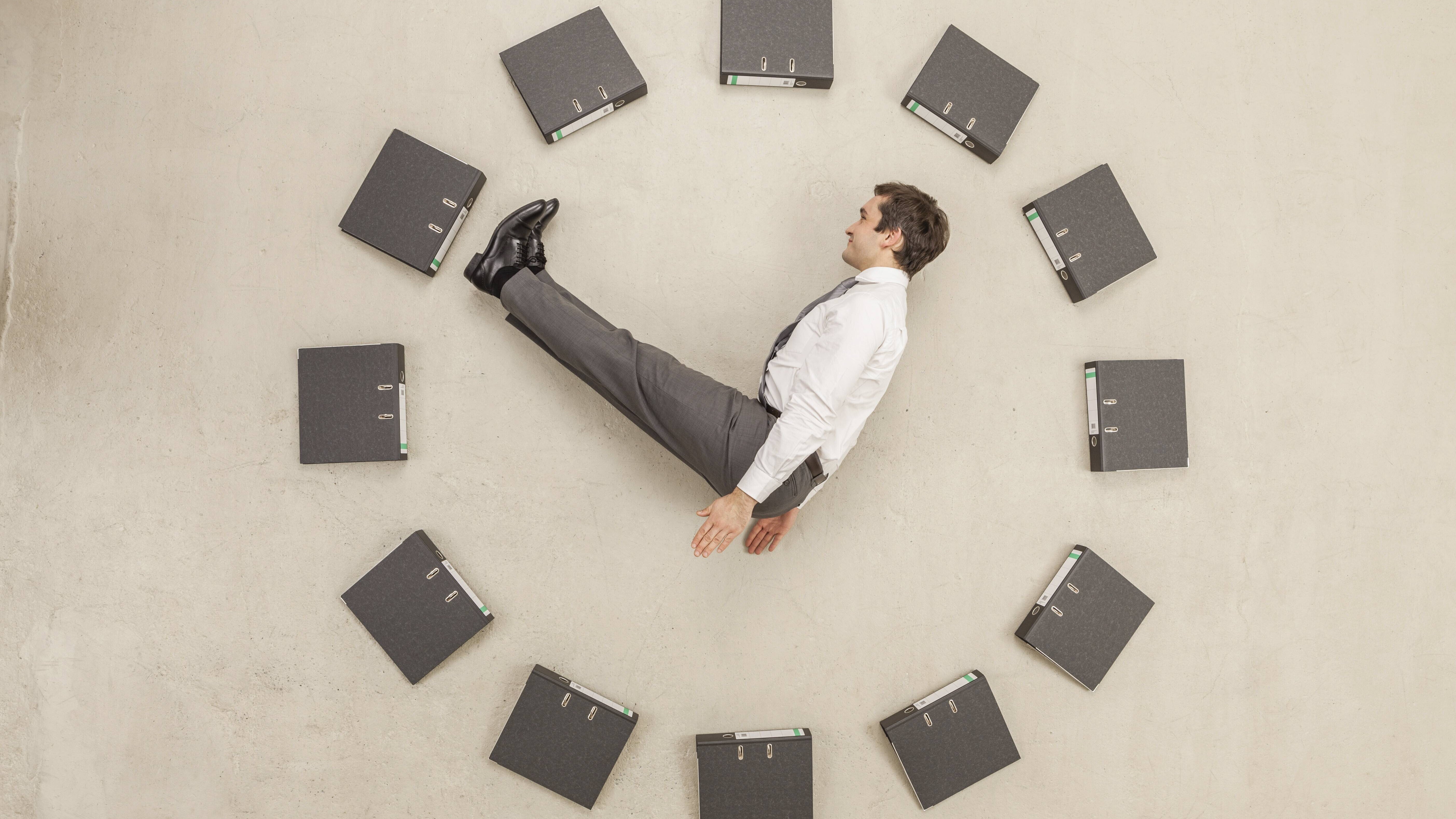 Aktenordner - kreisförmig auf dem Boden angeordnet - bilden eine Uhr. Ein Mann in Anzug stellt die Zeiger dar.