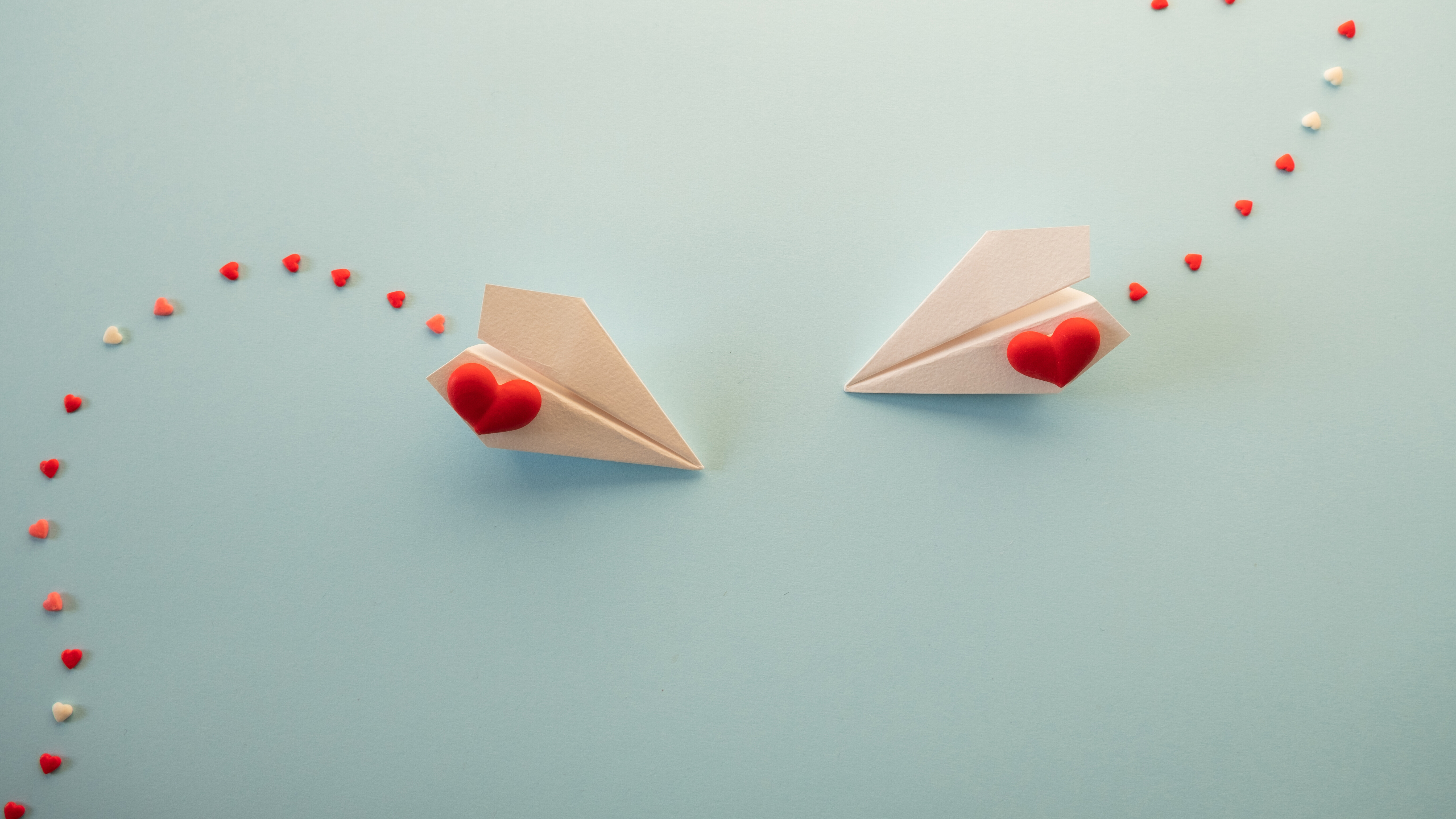 Zwei Papierflieger mit einem Herz fliegen aufeinander zu. Sie ziehen eine Spur von Herzen hinter sich her.