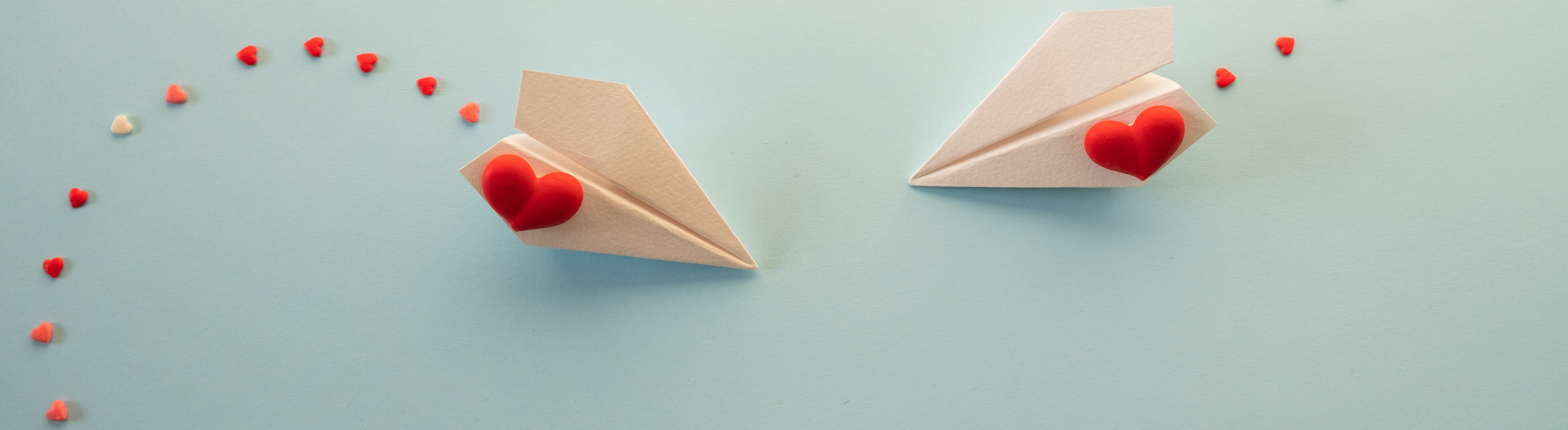 Zwei Papierflieger mit einem Herz fliegen aufeinander zu. Sie ziehen eine Spur von Herzen hinter sich her.