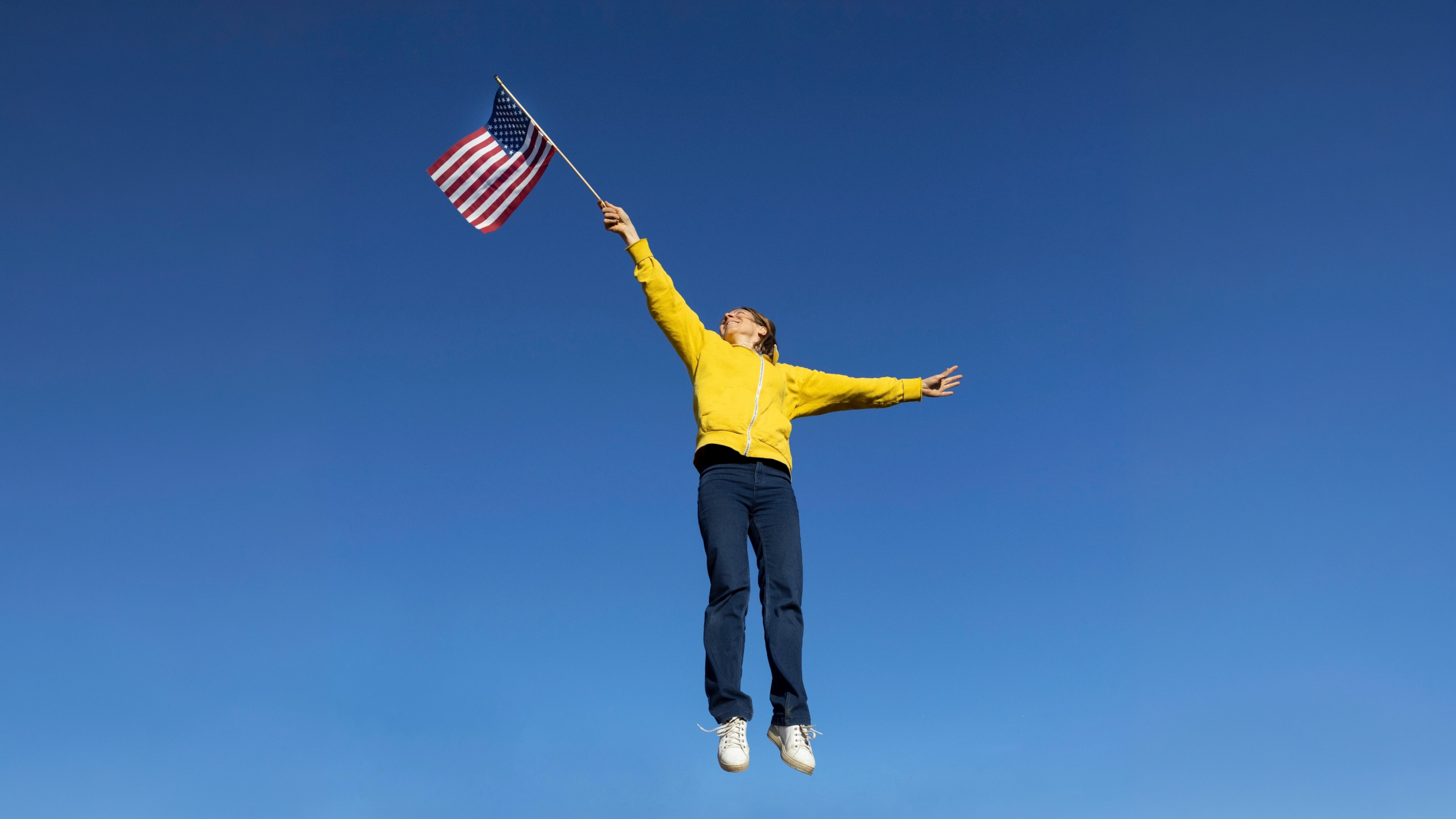 Eine Frau trägt ein gelbes Sweatshirt, eine dunkle Jeans und hält die amerikanische Fahne hoch mit einem ausgestreckten Arm und springt dabei in die Luft vor einem blauen Himmel.