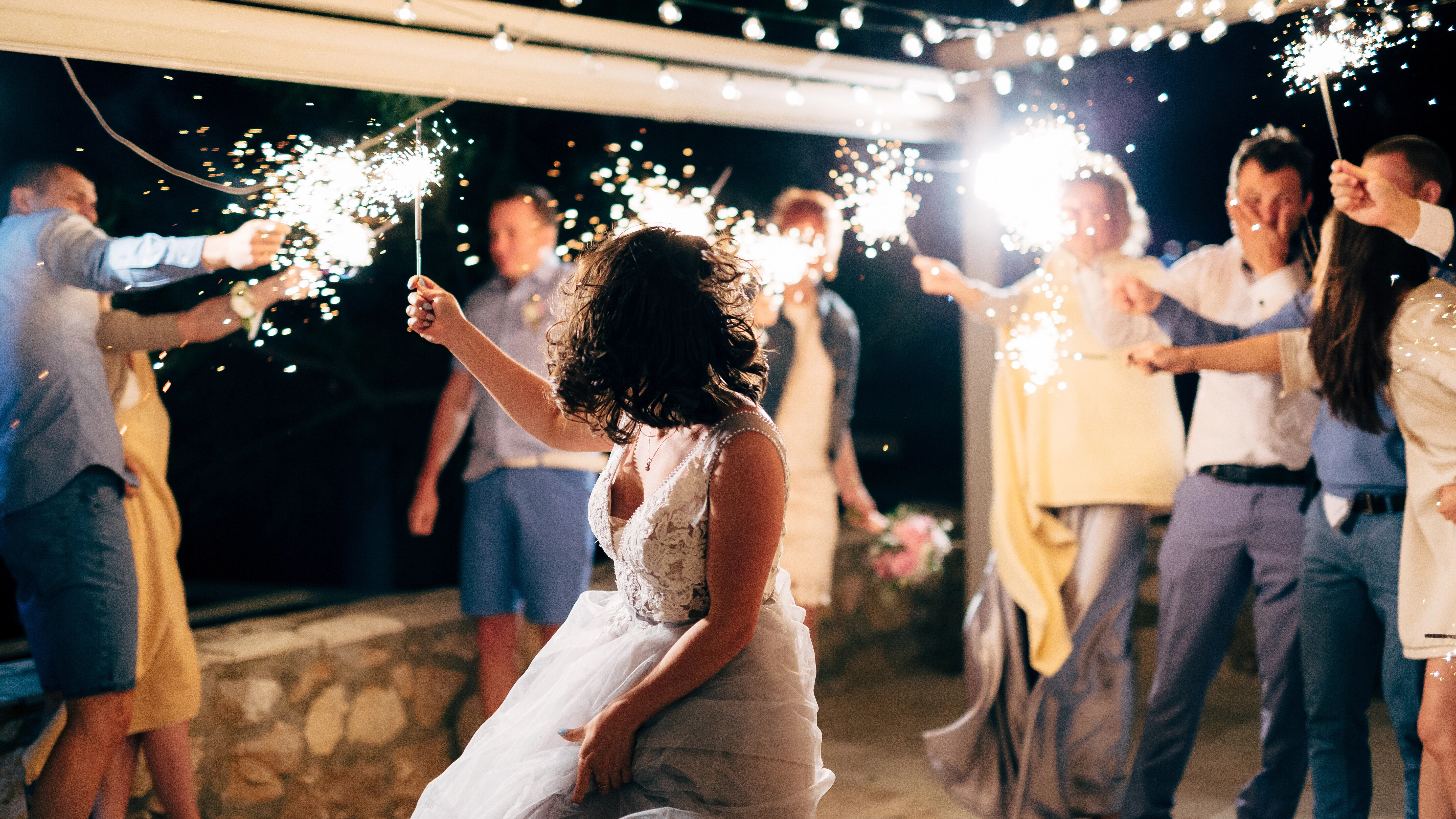 Eine Gruppe von Hochzeitsgästen tanzt bei Nacht im Kreis mit Wunderkerzen in der Hand. Eine Frau trägt ein Hochzeitskleid.
