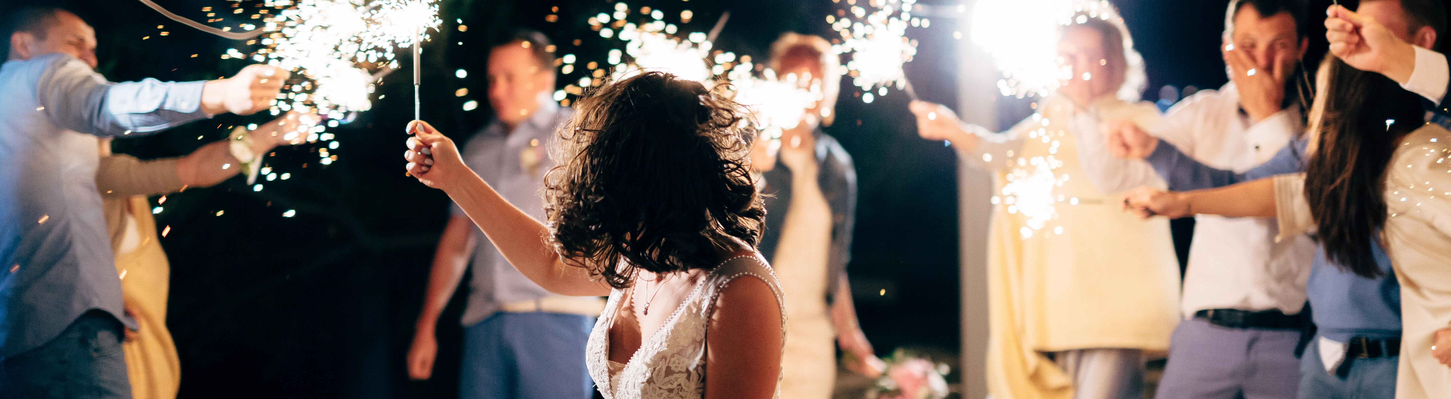 Eine Gruppe von Hochzeitsgästen tanzt bei Nacht im Kreis mit Wunderkerzen in der Hand. Eine Frau trägt ein Hochzeitskleid.