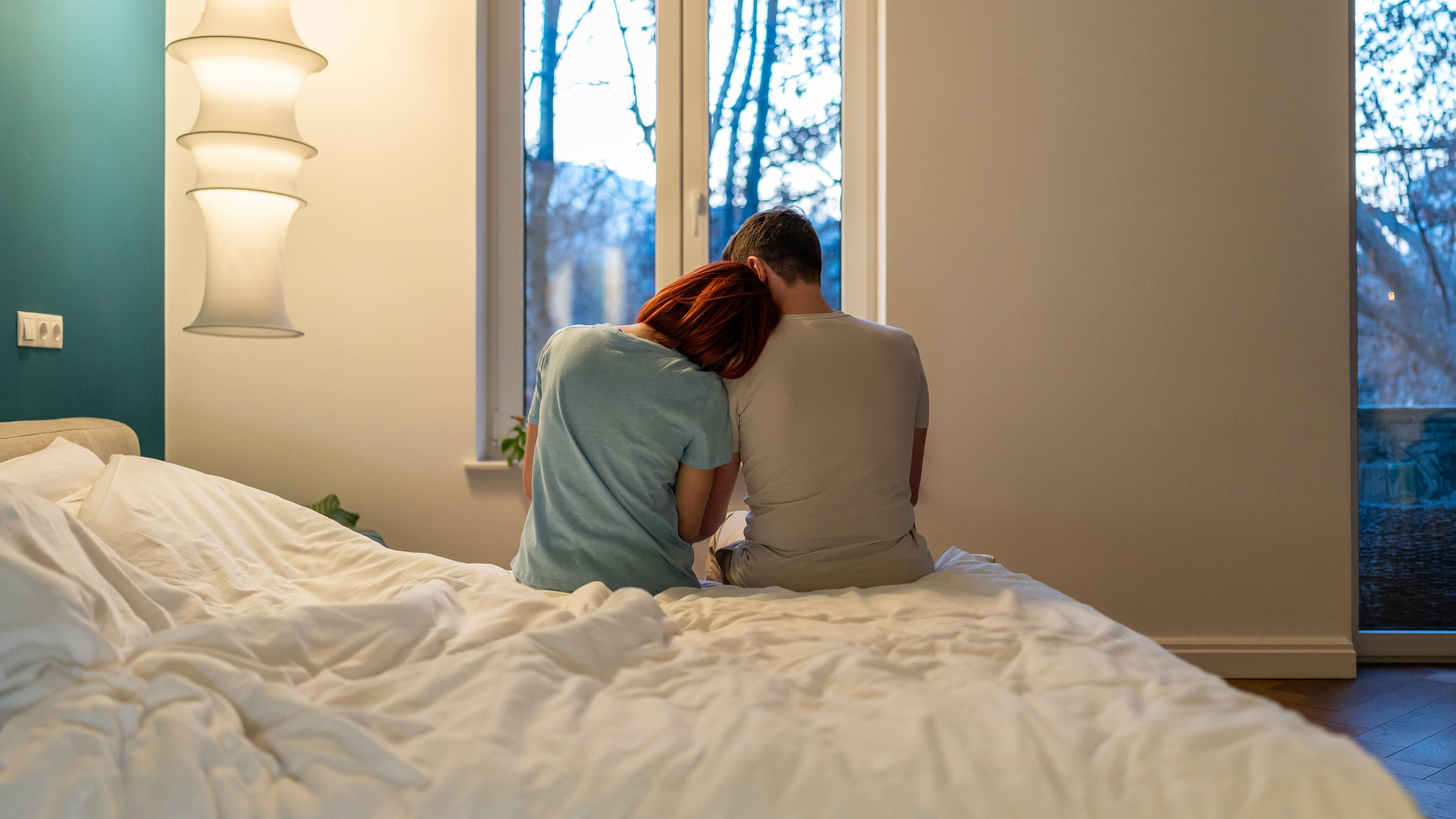 Eine Frau und ein Mann sitzen nebeneinander am Bettrand. Sie lehnt ihren Kopf an seiner Schulter an.