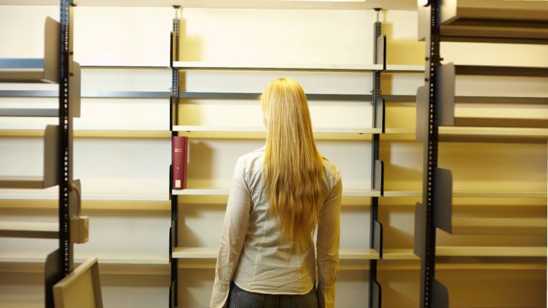 Studentin steht vor einem leeren Regal in einer Bibliothek und schaut auf das einzige Buch, das noch vorhanden ist.