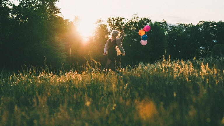 Eine Frau läuft auf einer Wiese. Hinter sich her zieht sie bunte Ballons. Die Sonne scheint. Es ist Abend.
