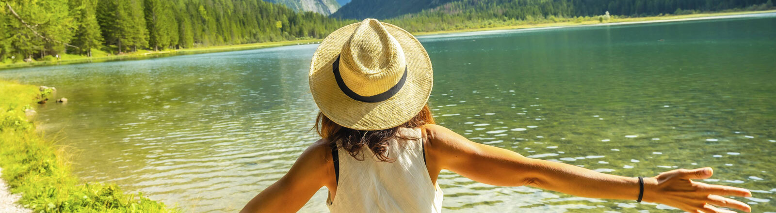 Frau mit Strohhut und offenen Armen genießt die unberührten Gewässer des Toblacher Sees und die majestätischen Dolomiten im Sommer