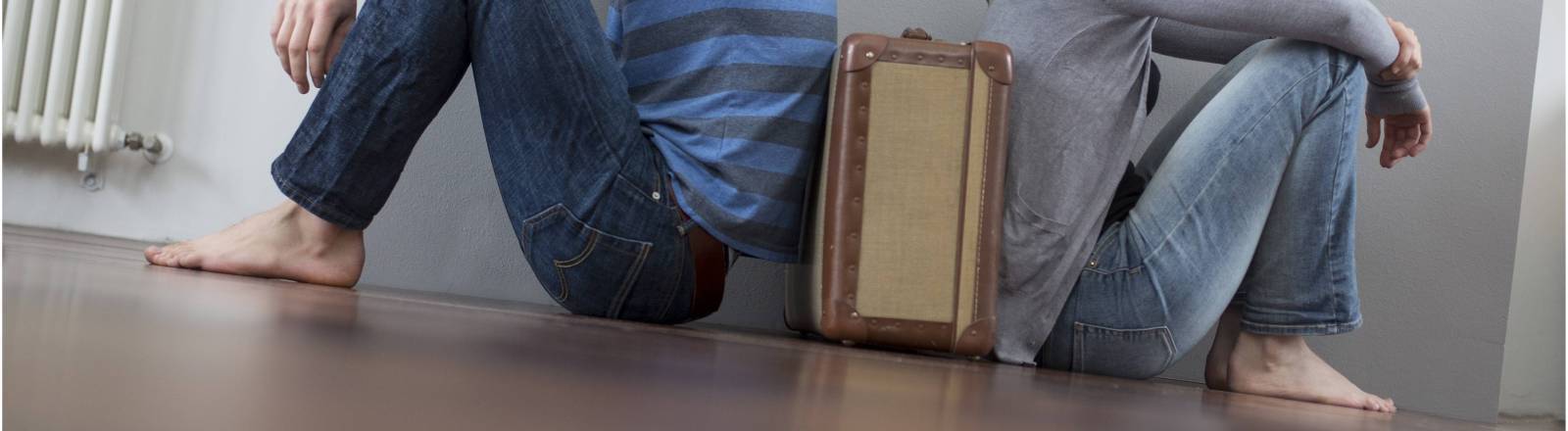 Mann und Frau sitzen mit dem Rücken zueinander auf dem Boden, zwischen ihnen steht ein Koffer