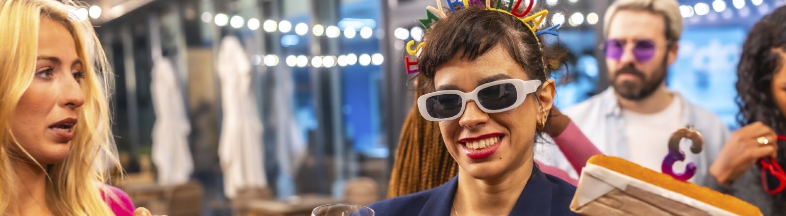 Eine Frau hält einen Kuchen in der Hand, in dem eine Kerze mit der Nummer drei steckt. In der anderen Hand hält sie ein Glas mit Bier. Sie trägt eine Sonnenbrille und einen Haarreif mit dem Schriftzug "It's my birthday".