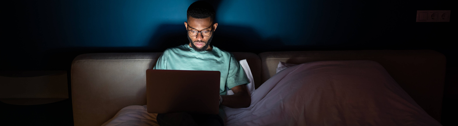 Junger Mann arbeitet an einem Laptop, liegt nachts im Bett und schaut aufmerksam auf den Bildschirm.