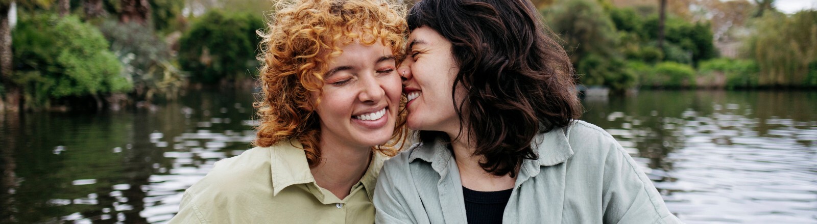 Zwei Frauen sitzen zusammen in einem kleinen Boot auf einem See. Eine Frau gibt der anderen einen inningen Kuss auf die Wange. Beide lächeln.