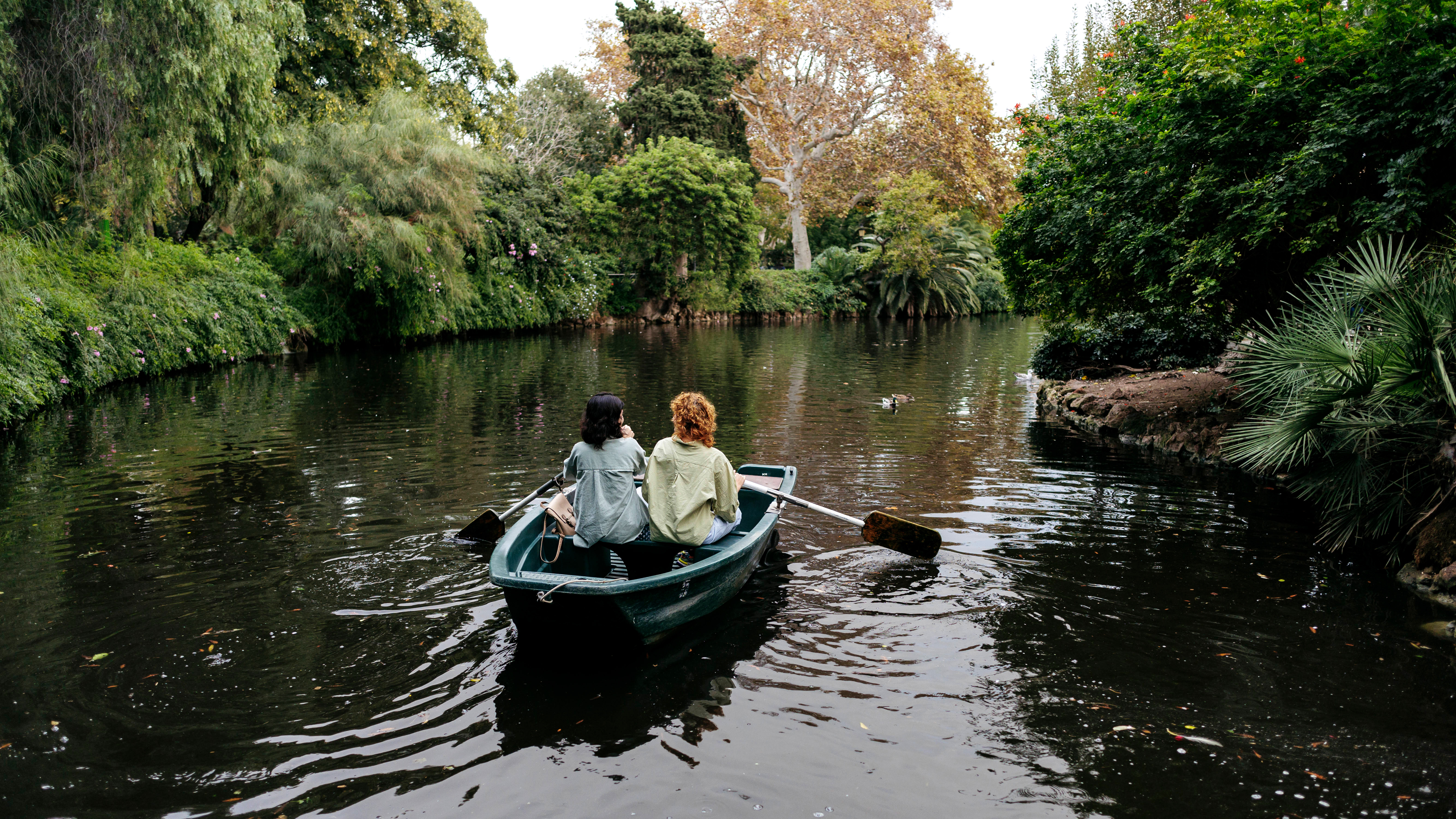 Lesbisches Pärchen in einem Ruderboot auf einem Weiher in Barcelona. Ringsum viel Grün.