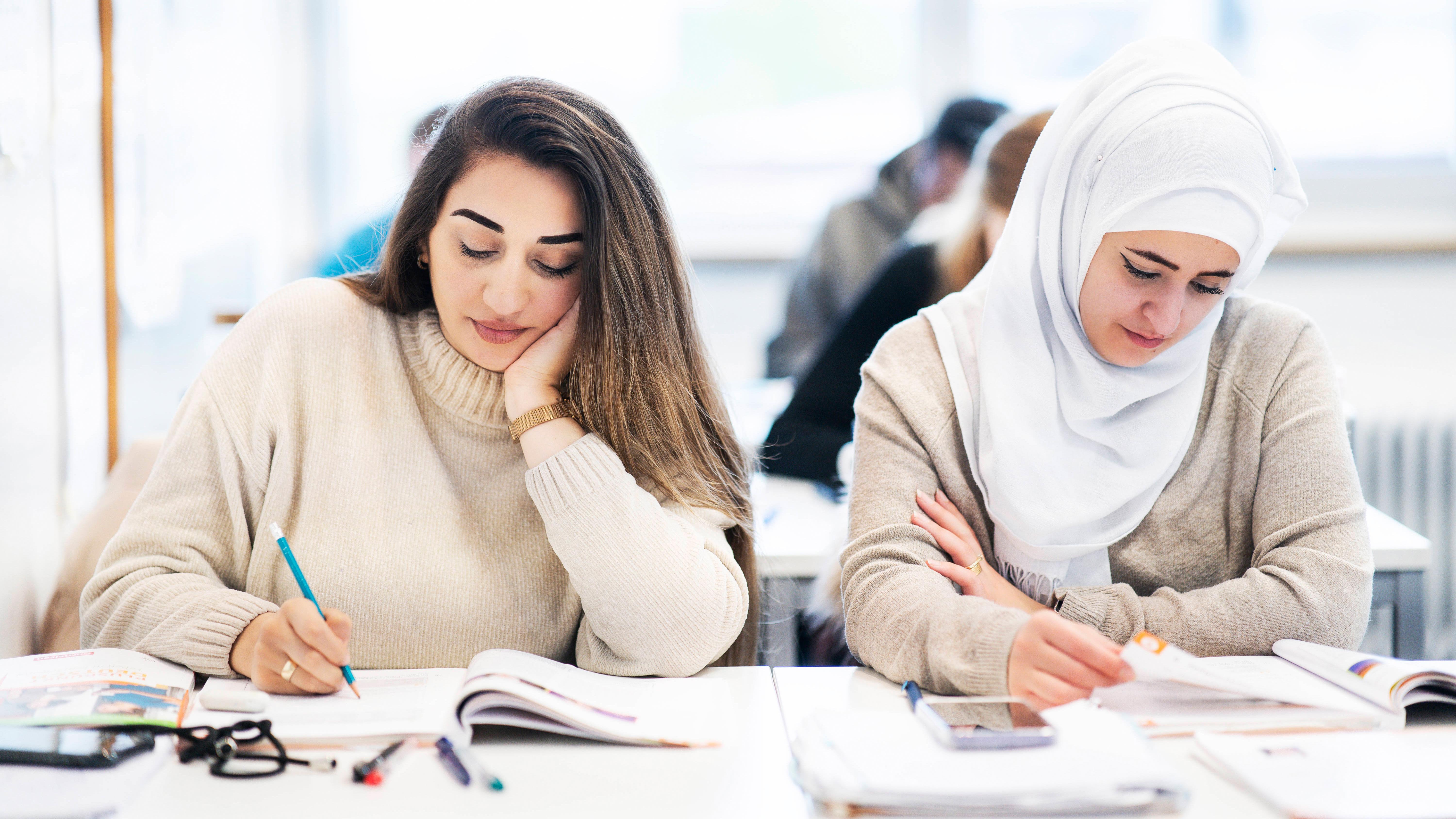 Zwei junge Frauen, eine von ihnen mit Kopftuch in einem Klassenzimmer schreiben etwas in ein Heft.