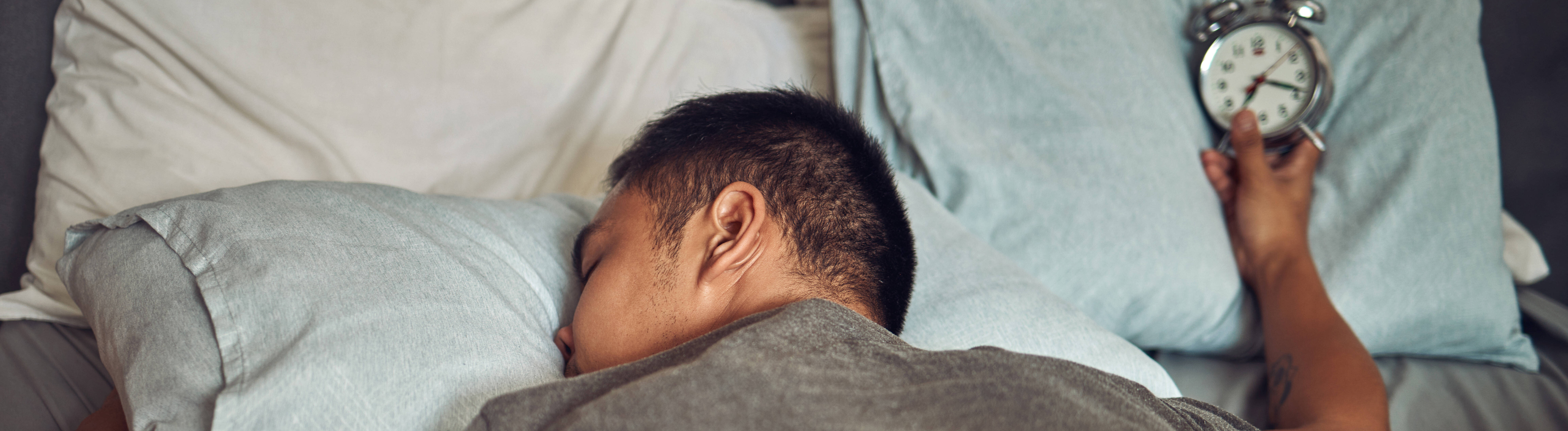 Mann mit Wecker liegt platt im Bett