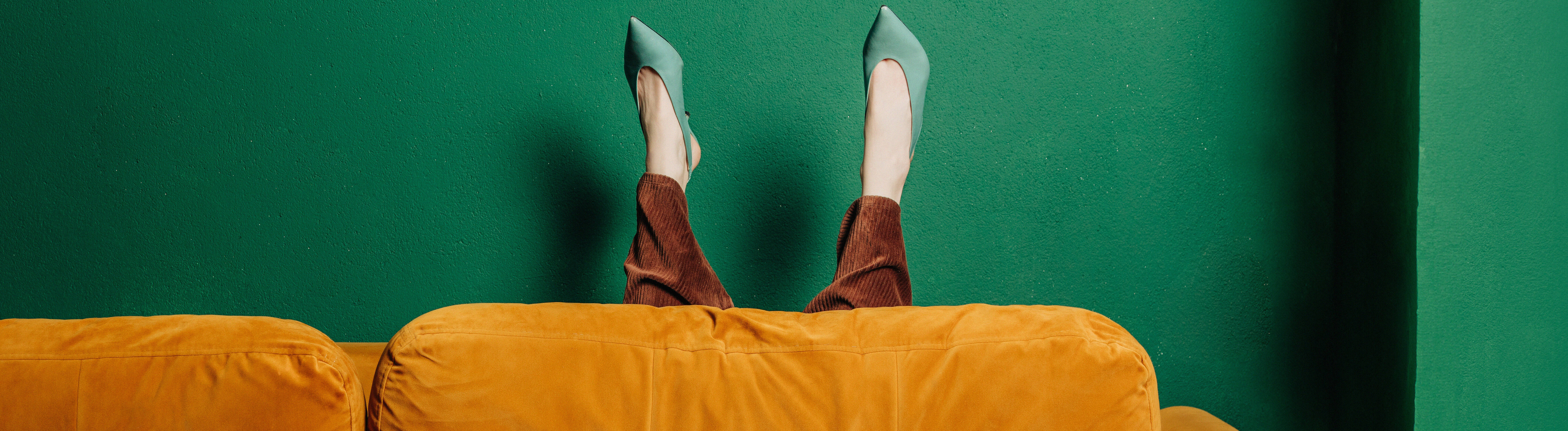 Die Beine einer Frau, mit grünen Absatzschuhen und einer braunen Hose, ragen hinter einem orangefarbenen Sofa hervor, die Wand dahinter ist grün
