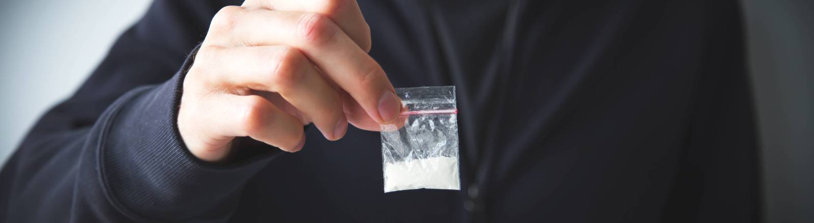 Person in Sweatshirt und Kapuzenpulli hält kleine Tüte mit weißer Substanz