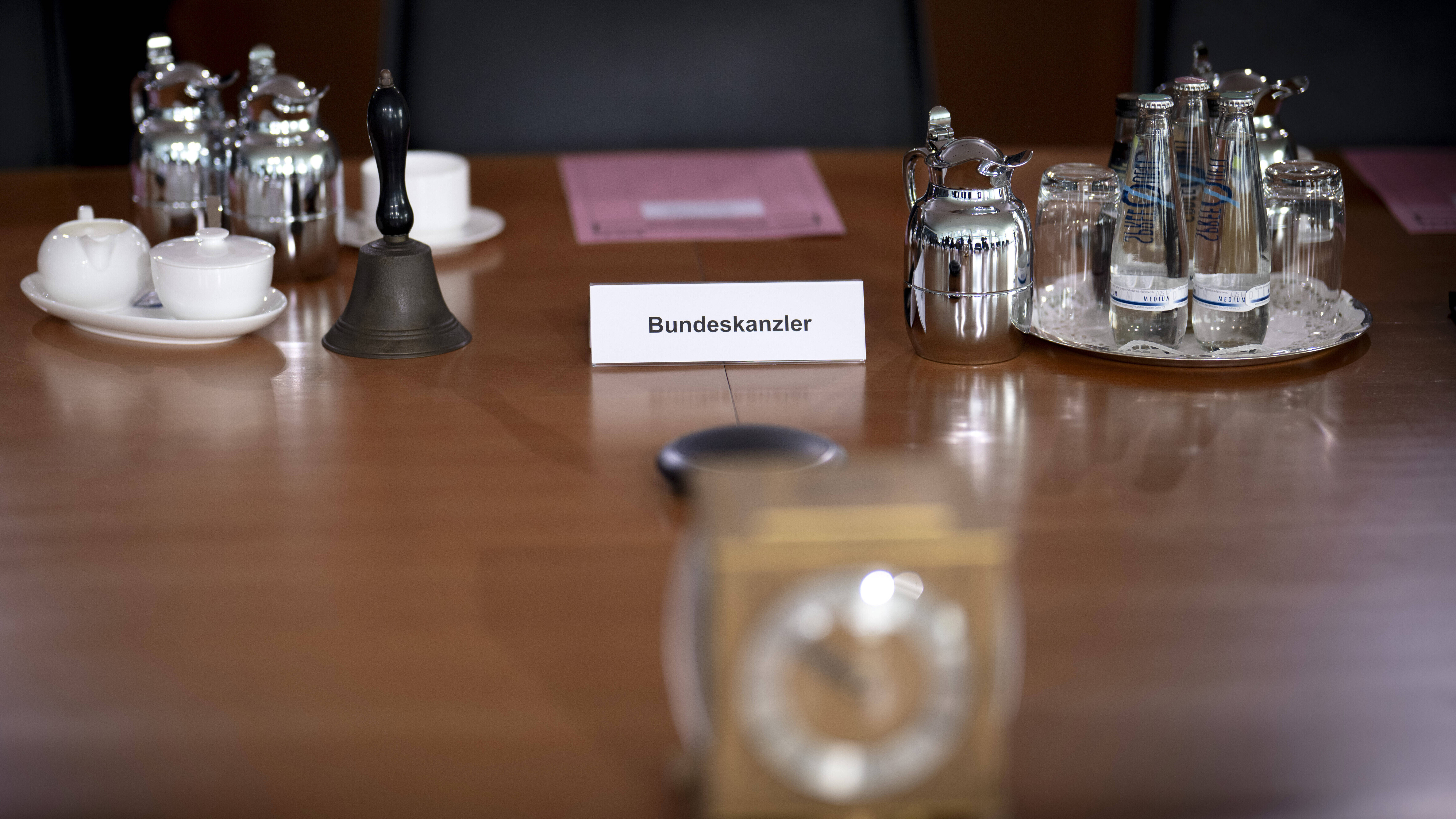 Ein Holztisch im Kanzleramt ist gedeckt mit Tassen, Gläsern, Wasserfläschchen und kleinen Kannen. Auf dem Tisch steht ein Aufsteller mit "Bundeskanzler".