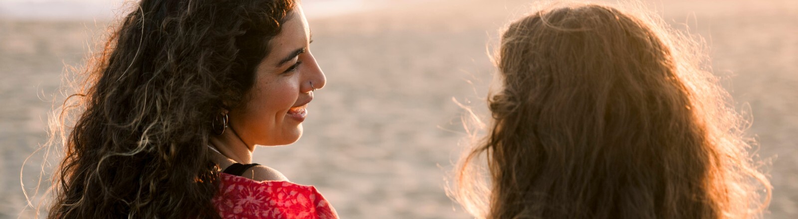 Zwei Frauen sitzen am Strand und unterhalten sich. Wir sehen sie von hinten.