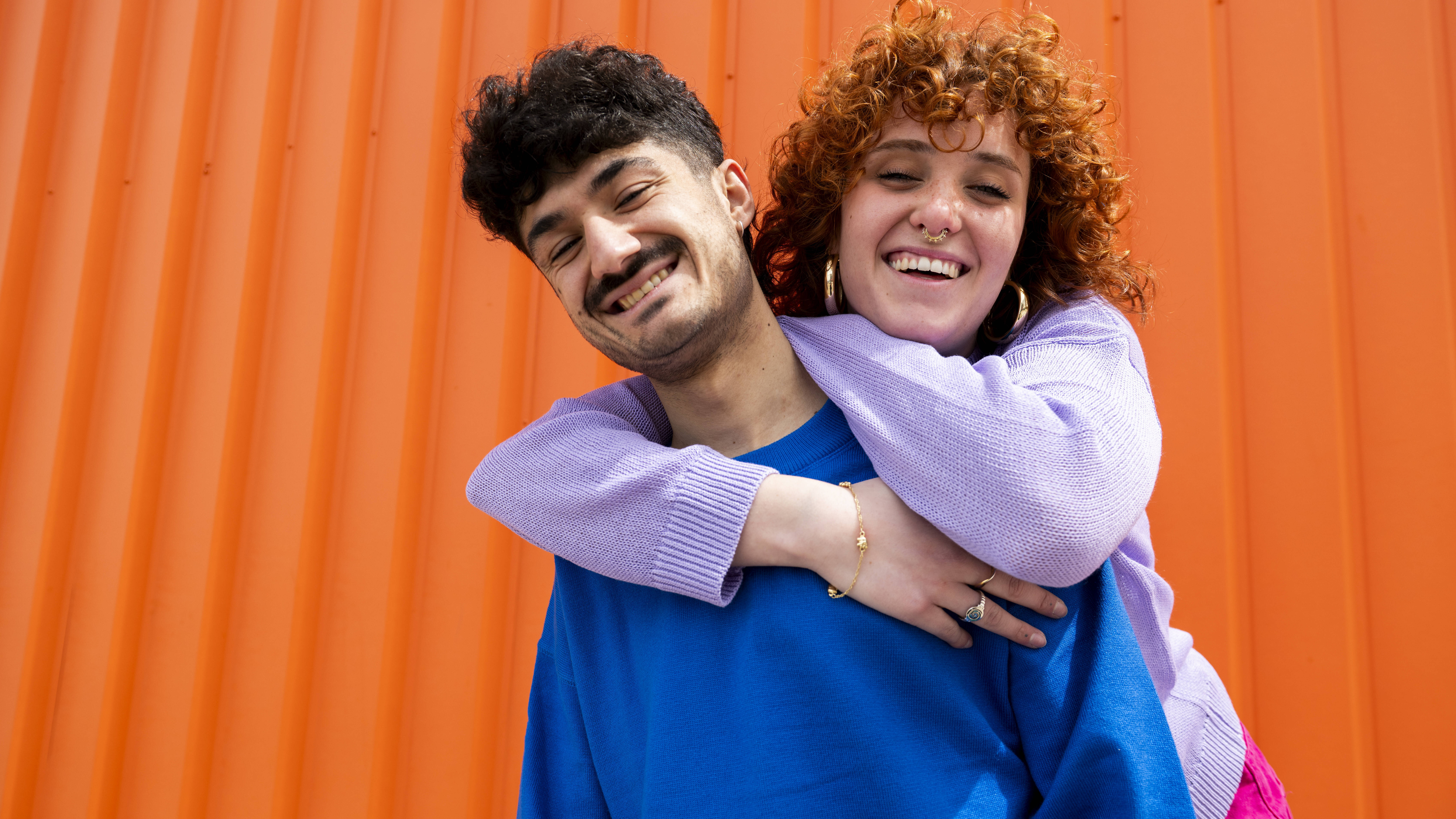 Eine Frau umarmt einen Freund von hinten. Die beiden stehen vor einem orangefarbenen Container