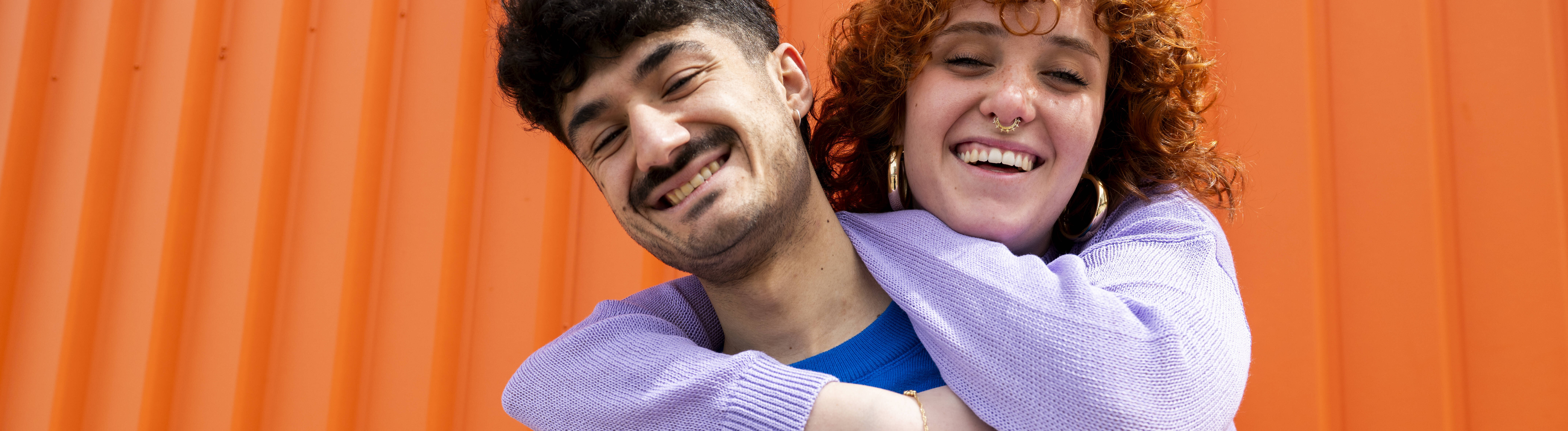 Eine Frau umarmt einen Freund von hinten. Die beiden stehen vor einem orangefarbenen Container