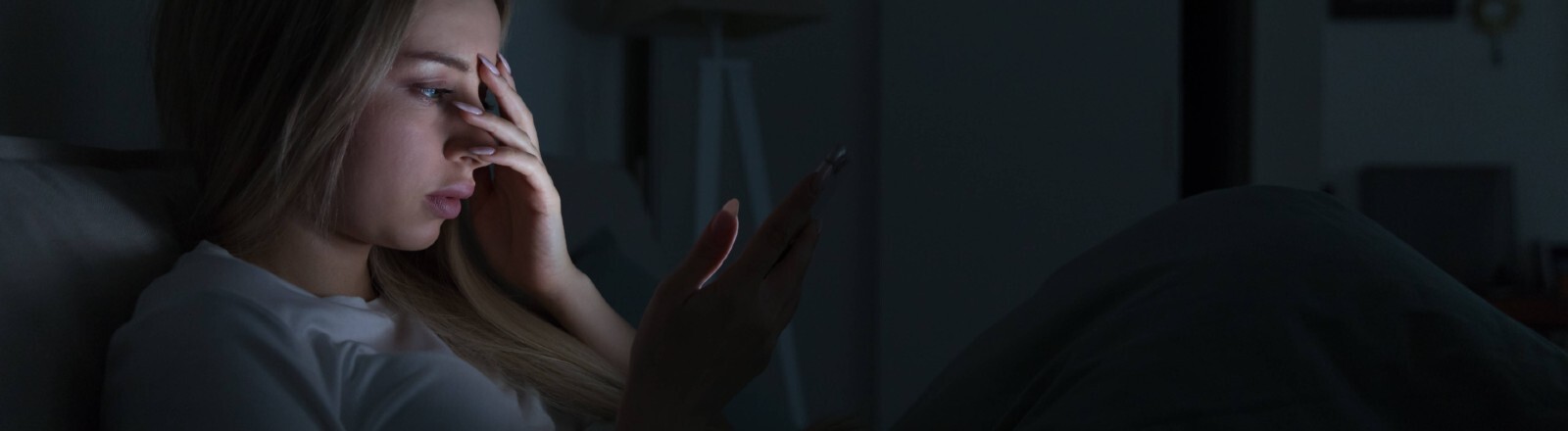 Eine junge Frau liegt in einem dunklen Zimmer in einem Bett. Das Licht ihres Handydisplays erhellt ihr Gesicht. Sie hält sich eine Hand vor das Gesicht.